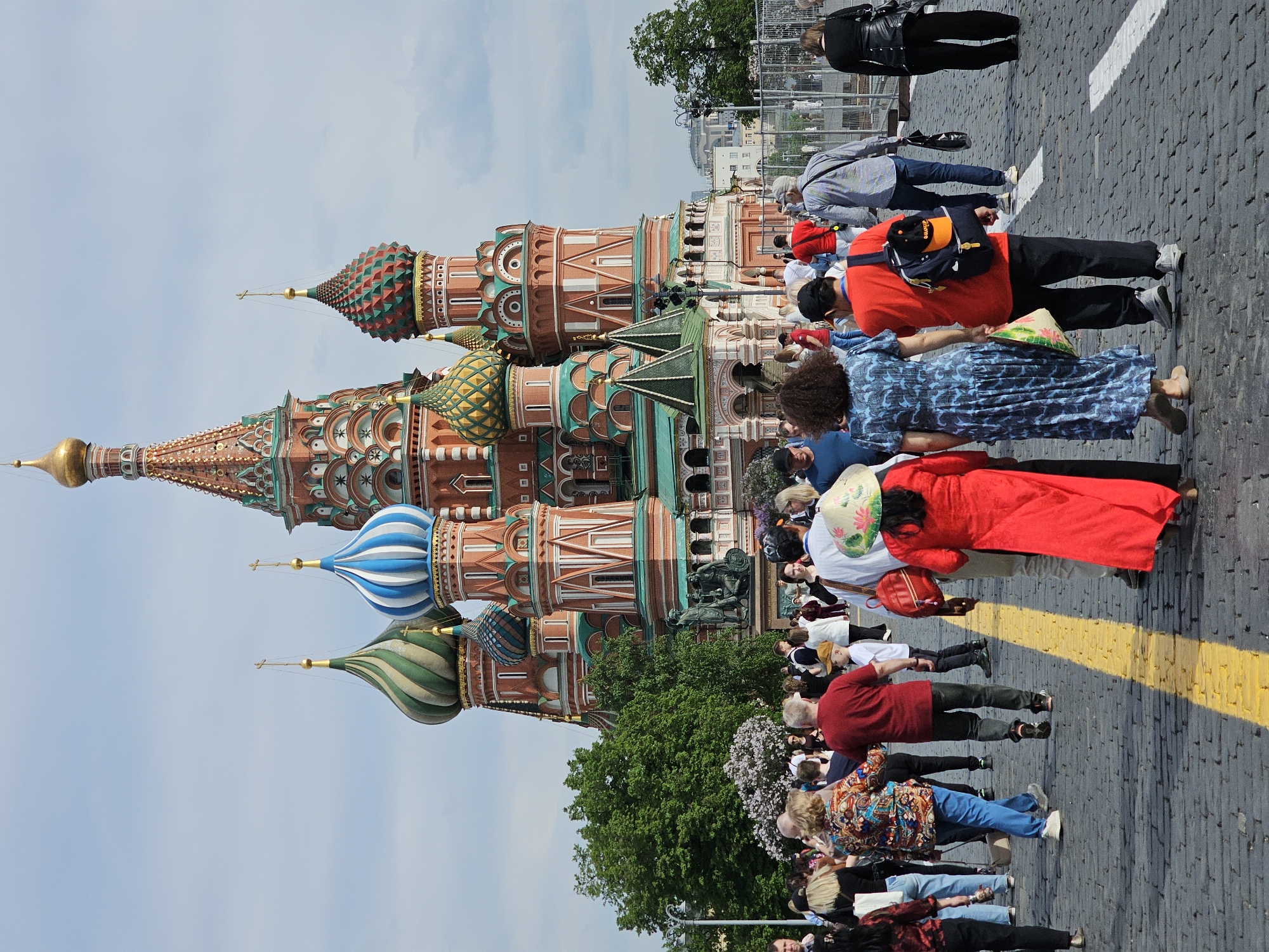 The image shows a crowd of people in front of the Saint Basil's Cathedral in Moscow, Russia. The cathedral is a prominent landmark with colorful onion domes and intricate brickwork. The crowd consists of tourists and locals, dressed in a variety of clothing. The ground is paved with cobblestones. The sky is partly cloudy.