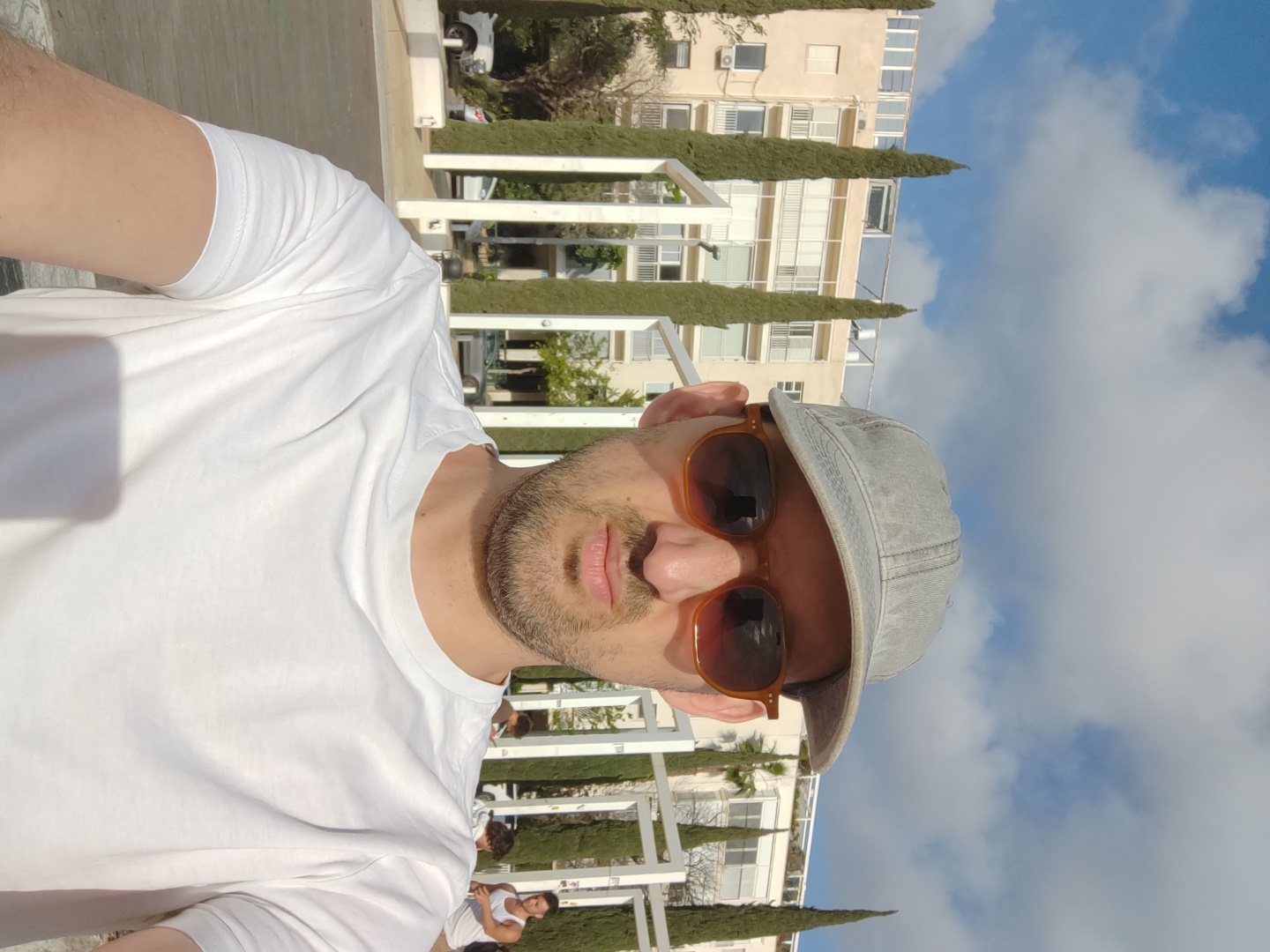 A man in a white t-shirt, grey cap, and sunglasses takes a selfie. He is outdoors in Tel Aviv, Israel. The backdrop features white pergola-like structures, tall, thin green trees, and a multi-story building under a blue sky with scattered clouds. People are visible in the background, engaging in activities near the white structures.