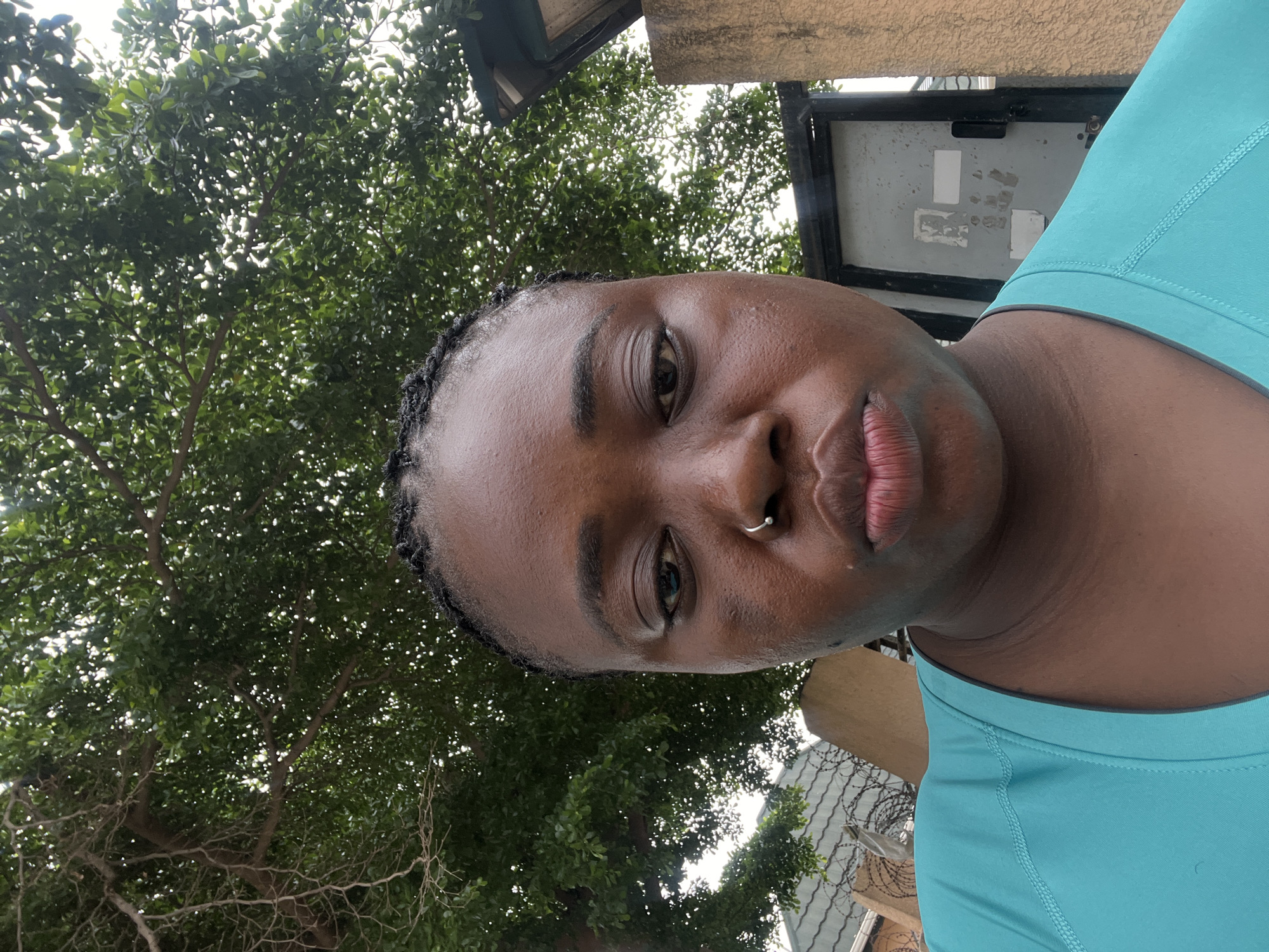 A medium shot portrait of a young Black woman is captured from a low angle, looking up at the camera. She has her braided hair pulled back and wears a teal-colored t-shirt. She has a nose ring in her left nostril. The background consists of lush green trees with dappled sunlight filtering through, indicating an outdoor setting. To the right of the woman, a concrete wall and a metal gate are visible, suggesting an urban or semi-urban environment. The presence of barbed wire on a fence in the lower left corner hints at security measures. The lighting suggests it is daytime, possibly overcast or in the shade of the trees. The scene's details, such as the type of trees and architectural elements, are consistent with common residential or public spaces in Abuja, Nigeria.