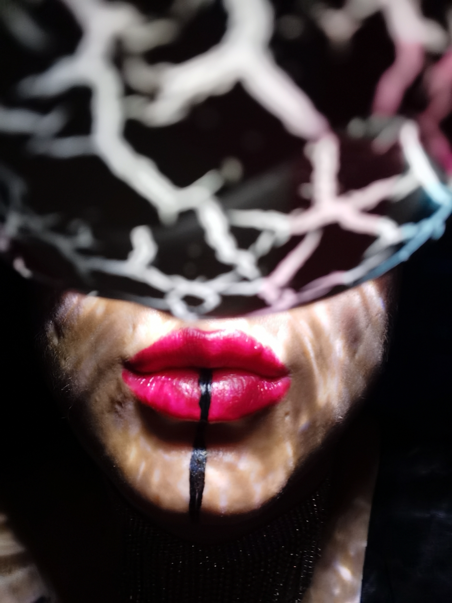 An extreme close-up photograph focuses on a person's lips, partially obscured by a patterned shadow reminiscent of cracked earth or branches against a dark background. The lips are painted a bright, glossy red. A thick, black line is drawn from the center of the lower lip downward onto the chin. The skin around the mouth is lit by dappled light, creating a mottled appearance. The upper portion of the image is dominated by the out-of-focus pattern casting the shadows. The lower portion of the frame fades into darkness. There is no indication of Chaikovsky, Russia.