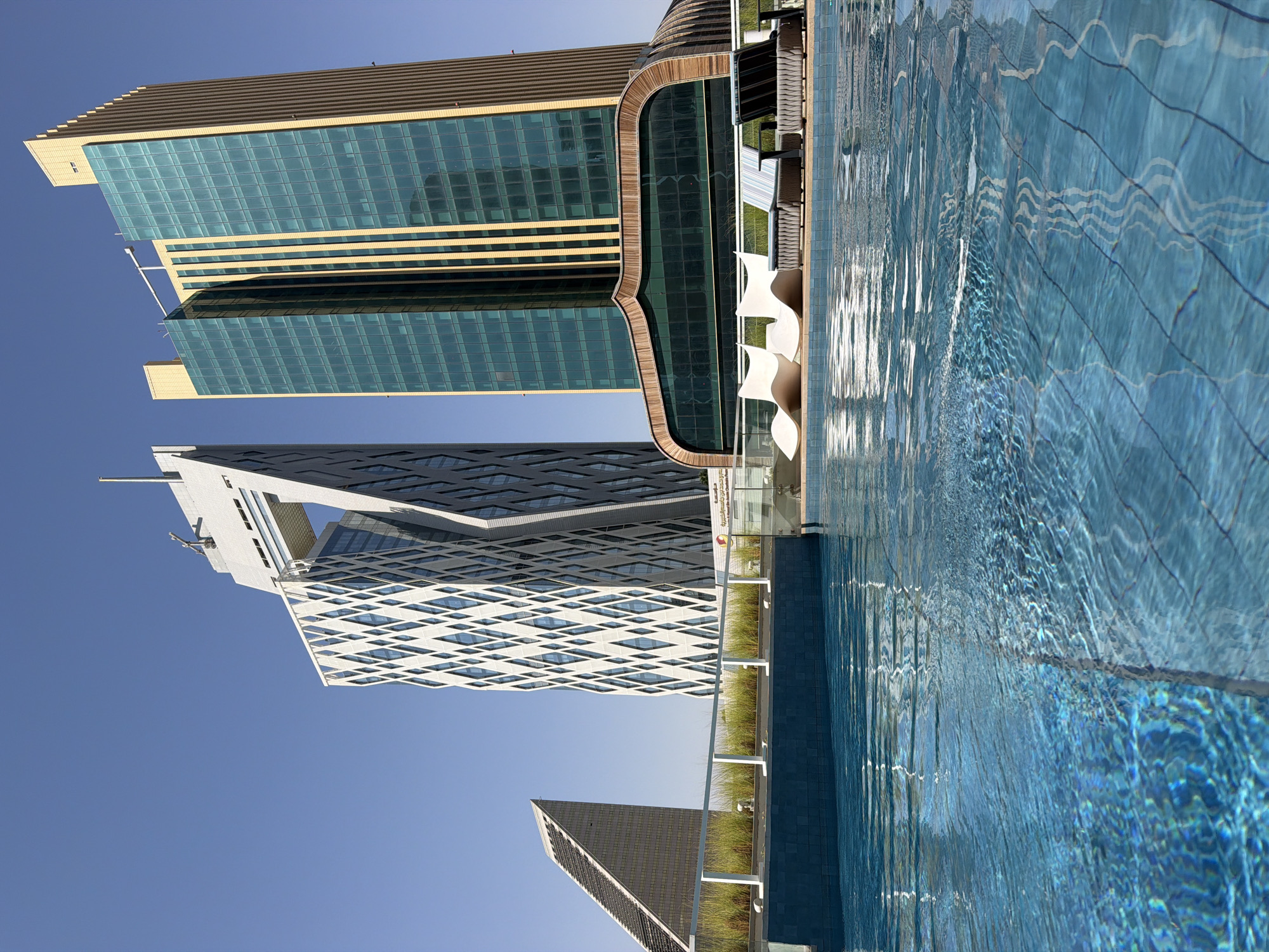 This is a wide, eye-level, outdoor shot of a swimming pool in the foreground, with a city skyline in the background. The pool water is a bright blue and reflects the sky and the surrounding architecture. There are three lounge chairs and a small table at the edge of the pool. Behind the pool is a white railing and a row of tall green grass. In the background, there are three modern skyscrapers. The leftmost building has a unique, geometric design with diamond-shaped patterns. The middle building is a tall, rectangular glass skyscraper with a golden facade. The rightmost building is also a tall, rectangular glass skyscraper. The sky is clear and blue. The scene appears to be a resort or hotel setting.