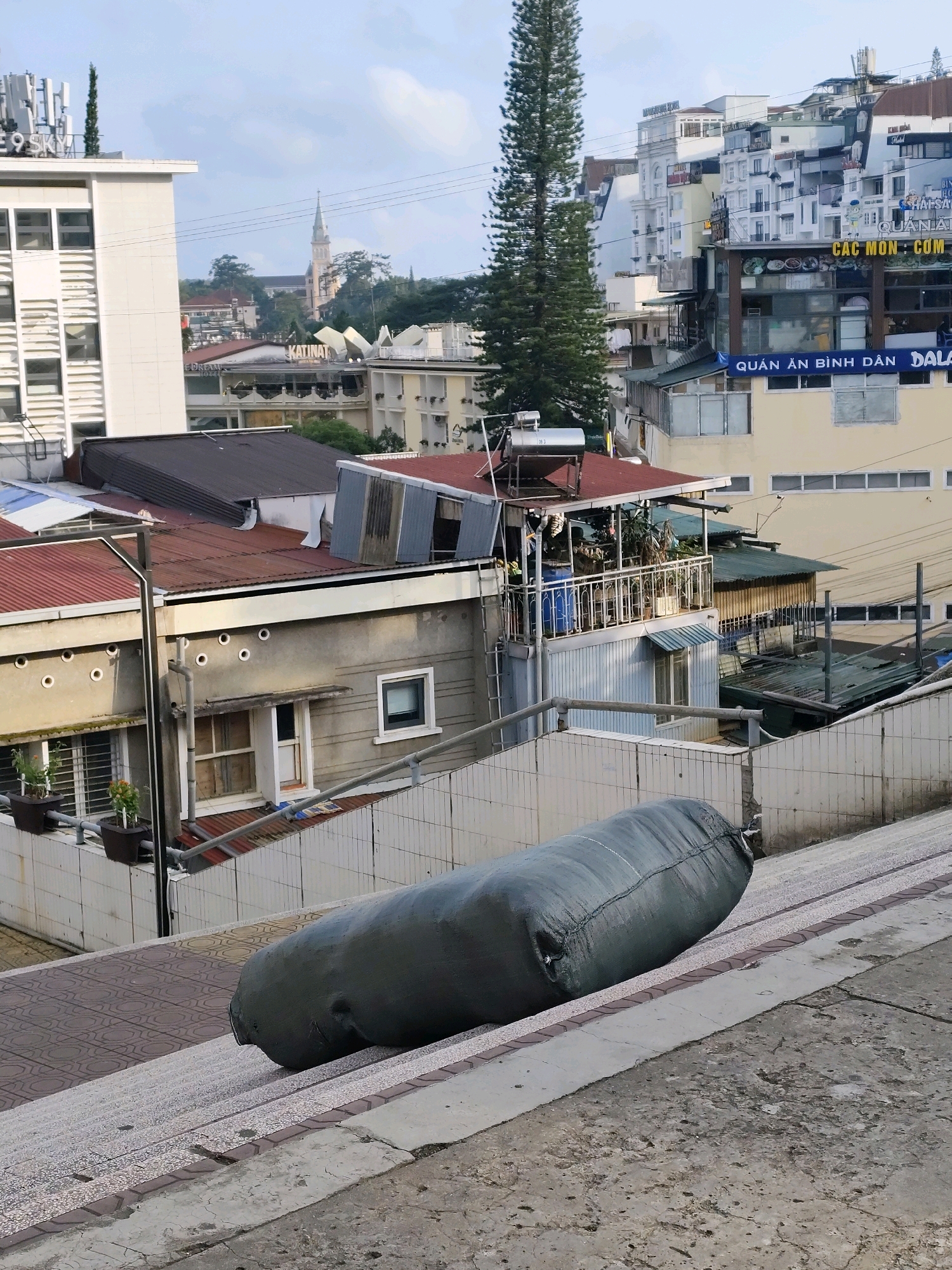 The image depicts an outdoor scene in Da Lat, Vietnam, likely from a higher vantage point looking down. In the foreground, a large, dark green, cylindrical object, possibly a sandbag or a large tube, rests on a set of angled stairs. The stairs are constructed with a mosaic-like pattern on the treads and a stone-like material on the risers. Beyond the stairs, a tiled wall runs horizontally, separating it from a residential or commercial area.
This area is densely populated with buildings of varying heights and architectural styles. Several rooftops are visible, many made of corrugated metal in shades of red or dark gray. Some buildings have balconies with railings and greenery. In the background, taller structures with multiple windows are prominent, indicative of apartments or hotels. Signs with Vietnamese text, such as "QUÁN ĂN BÌNH DÂN" (meaning "Budget Eatery") and "DALAT," are visible on some of the buildings, confirming the location. A tall, slender pine tree stands out against the sky, a common sight in Da Lat's landscape. A distant church steeple is also visible, suggesting a religious institution within the city. The sky is partly cloudy, casting a diffused light over the scene. There are no people directly visible in the frame, and the overall impression is one of a moderately built-up urban environment with some signs of infrastructure and everyday objects.