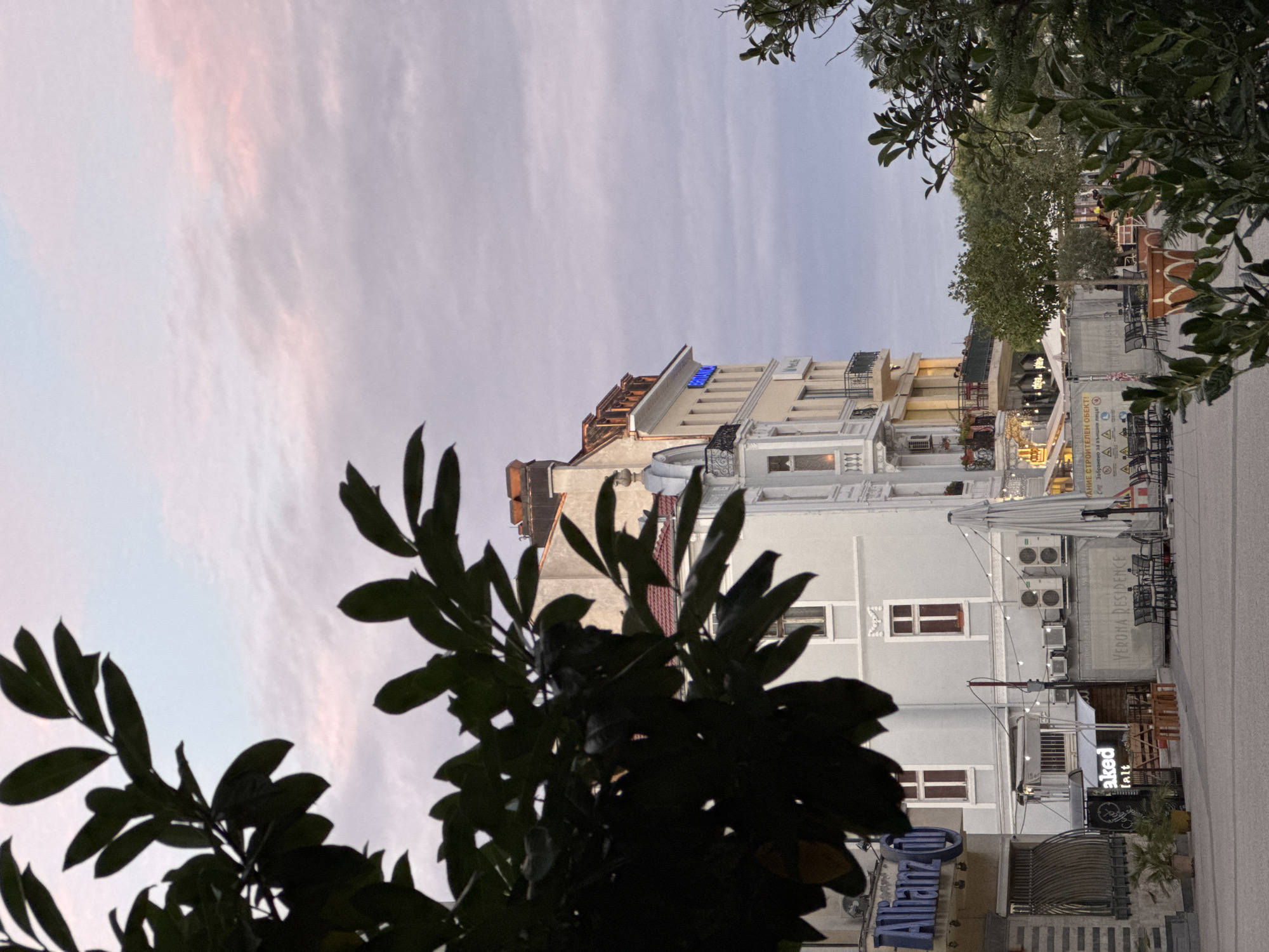 The image captures a street scene in Plovdiv, Bulgaria, likely in the pedestrian zone of the city center, with a prominent view of buildings and outdoor seating areas. In the foreground, dark green foliage from trees partially obscures the view. Beyond the foliage, the pastel-colored sky with light clouds suggests it is either early morning or late evening.
The buildings are distinct architectural styles. On the left, a building displays a blue "Allianz" logo. Adjacent to it is another building with light gray and cream facades, featuring multiple windows with red shutters. One building, a taller, yellow structure, has balconies on its upper floors and a blue illuminated sign that reads "BRIANCA".
Below these buildings, various outdoor seating areas are visible. Tables and chairs are arranged, some with umbrellas, suggesting cafes or restaurants. One area has a sign that reads "VERONA RESIDENCE". Further back, construction fencing is present with text in Bulgarian, indicating ongoing work. Several air conditioning units are mounted on the exterior walls of the buildings. A few potted plants are placed near the entrance of one establishment. The ground appears to be paved, typical of a city square or street. There are no clearly visible individuals engaging in specific actions, but some figures can be faintly discerned on the balconies of the yellow building.