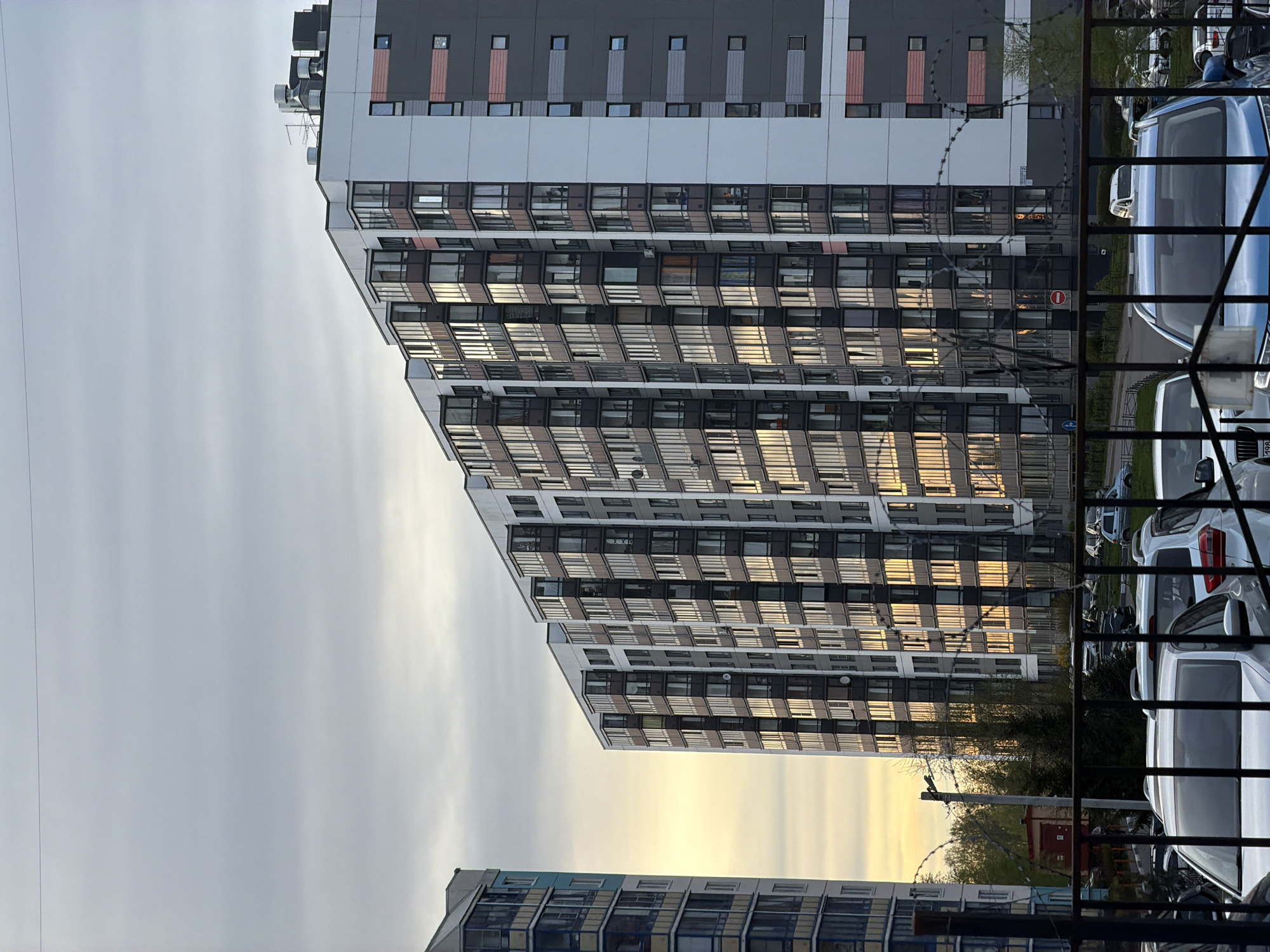 The image depicts a multi-story apartment building in Pavlovo, Russia. The building has a modern design with alternating grey and white sections and numerous windows that reflect the sunlight. In the foreground, there is a metal fence with barbed wire. Parked cars are visible behind the fence. The sky is overcast.