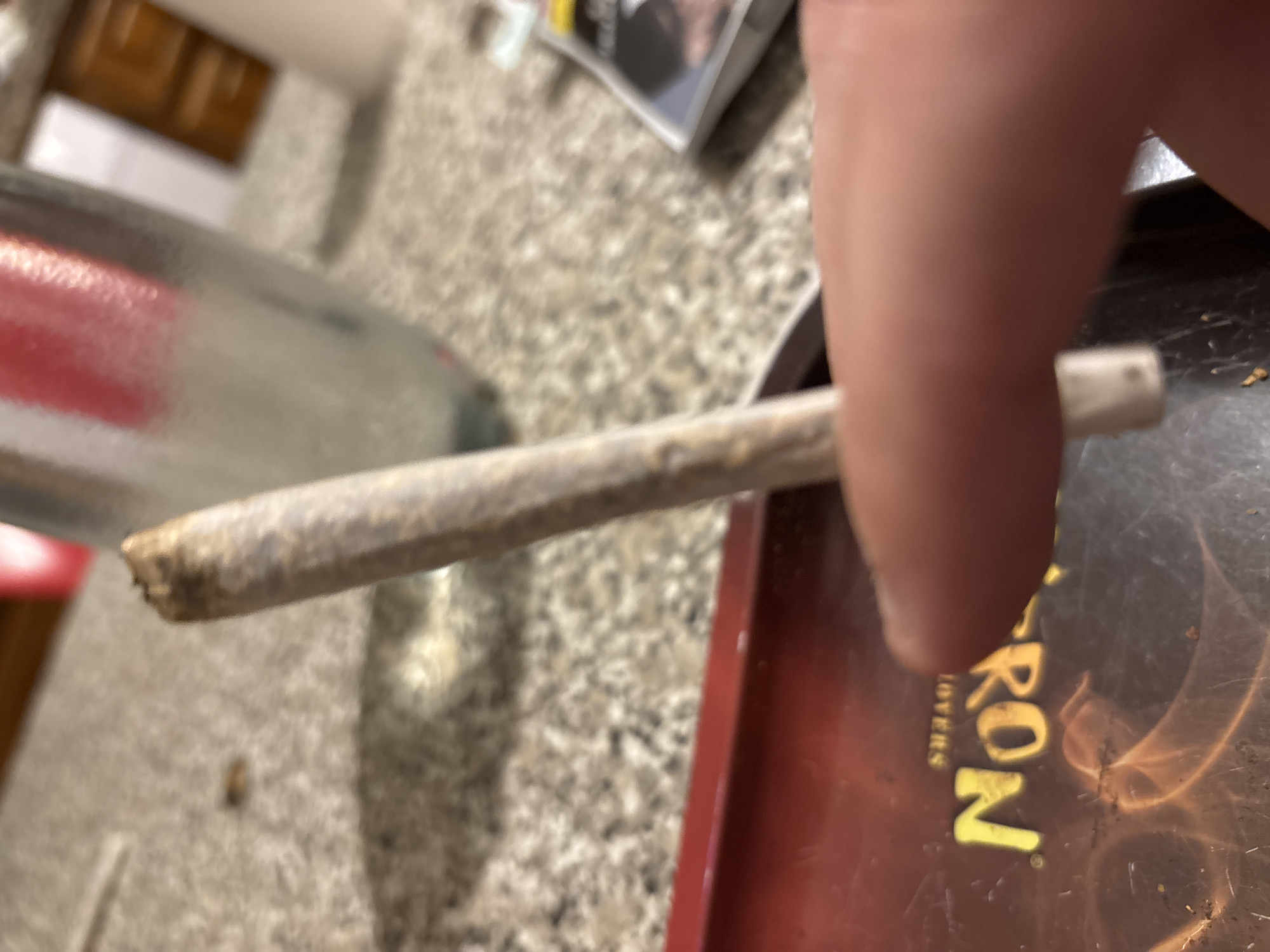 The image depicts a close-up, indoor scene with a primary focus on a hand holding a hand-rolled cigarette, commonly referred to as a "joint" or "spliff." The cigarette is light-colored, cylindrical, and shows a filter at one end, with visible plant material at the other end. A light-skinned thumb and index finger are visible, securing the cigarette above a reddish-brown rolling tray. The tray features yellow, inverted text reading "NORTON" and "LOVERS," along with a flame-like graphic and scattered specks of plant matter.
The background consists of a speckled, light-toned countertop or table surface. A tall, clear glass object, possibly a bottle, with a red internal element or reflection, stands behind the cigarette. Further in the background, blurred elements include a brown wooden cabinet or door and a small stack of papers or a magazine with white and yellow accents. The lighting is artificial, characteristic of an interior setting.
The location is specified as the city of Bragança, Portugal.