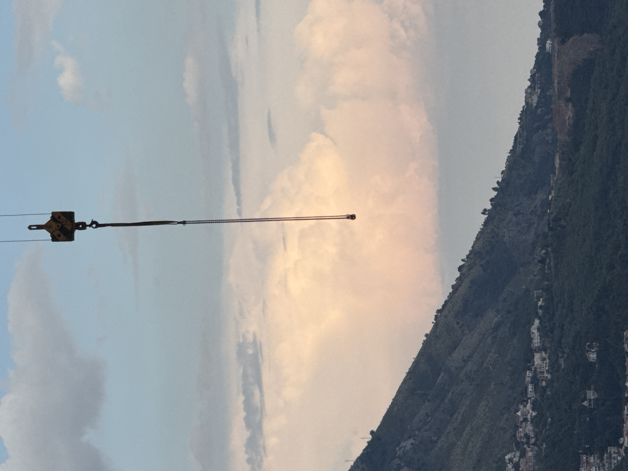 The image depicts a dramatic landscape with a focus on a crane and a mountainous terrain. A crane's hook and part of its lifting cable are visible in the left foreground, extending horizontally across the frame. The cable is taut, suggesting it is under tension, possibly lifting or holding a load, though the load itself is not clearly visible.
The background is dominated by a steep, densely forested mountainside, likely part of the Boreti area in Montenegro, given the context. Scattered white buildings are visible nestled within the foliage on the mountainside, indicating a populated area. The sky above is a blend of pale blue and soft, warm-toned clouds, suggesting it might be late afternoon or early morning. The light filtering through the clouds casts a gentle glow on the scene. The overall atmosphere is one of industrial activity juxtaposed with natural beauty. There is no visible text in the image.