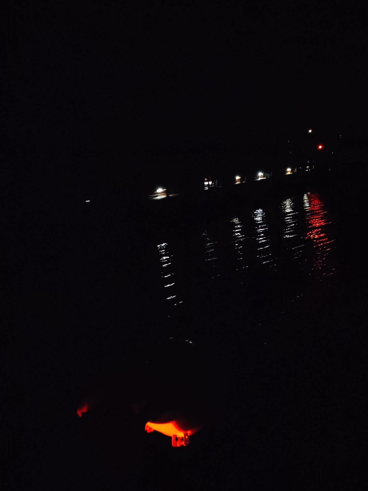 The image appears to be taken at night in Pruzhinino, Russia, near a body of water. There are lights visible across the water, including white and red lights which could indicate buildings or traffic. Reflections of these lights are visible on the water surface. In the foreground, there is a blurred orange glow, potentially from an object or source of light that is close to the camera. Due to the darkness and limited visibility, further details are difficult to determine with certainty.