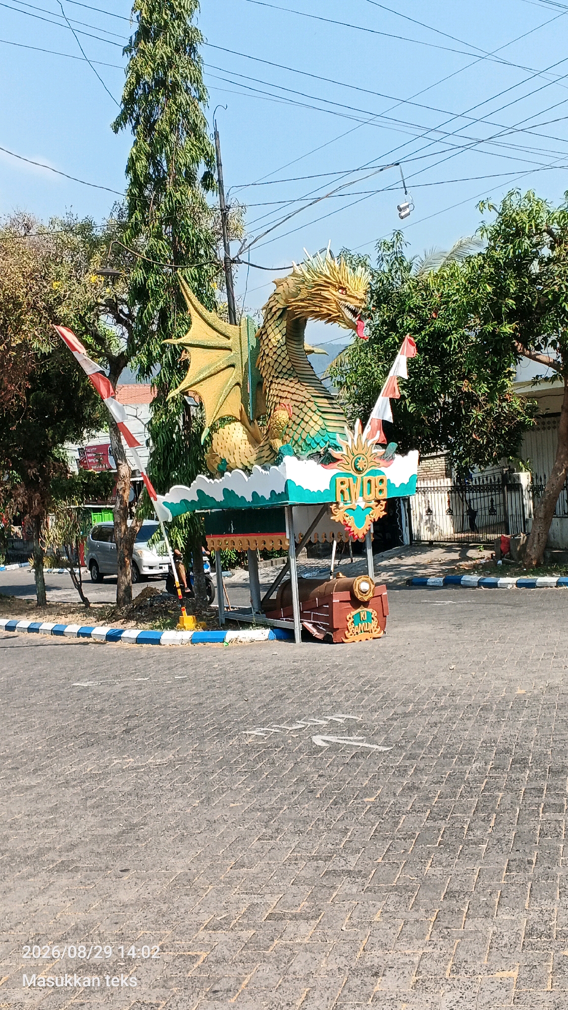 The image shows an outdoor scene on a sunny day. In the center is a decorative structure featuring a large, golden dragon statue perched atop a colorful platform. The platform is decorated with green, white, and red elements, including flags and an emblem that reads "RVO8". Below the platform, there is a model of a brown train car with a chest in front of it. The structure is situated on a paved street with white and blue striped markings along the edge. Trees and buildings are visible in the background, and power lines crisscross the sky. A silver minivan is parked on the left side of the street. The scene appears to be in a public area, possibly a street or intersection in the city of Sumberporong, Indonesia, given the timestamp and Indonesian text overlay in the lower left corner.
