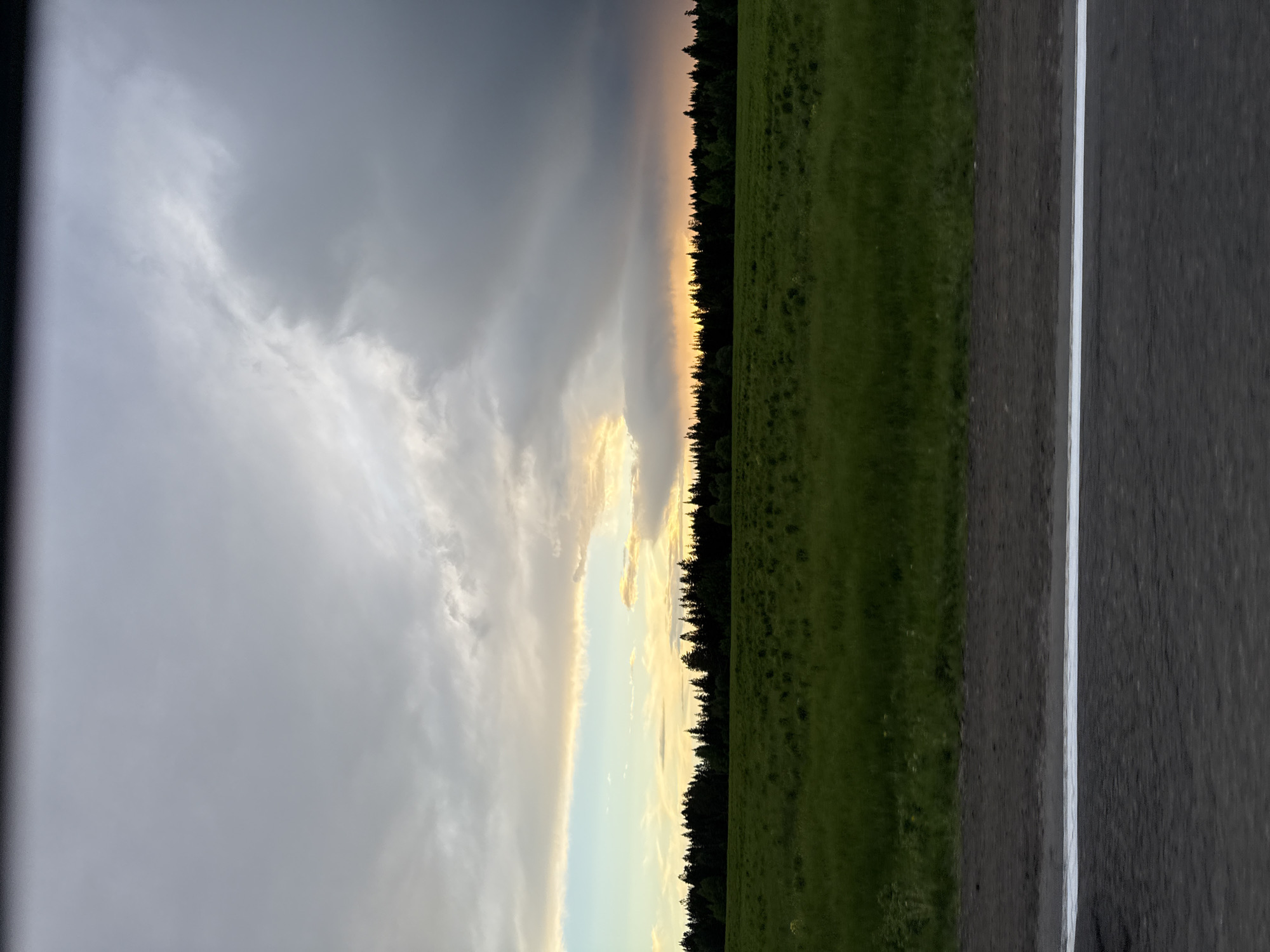 The image shows a landscape with a road in the foreground, followed by a green field, a line of trees, and a dramatic sky with dark storm clouds and a glimpse of golden light. The road has a white line marking its edge. There are no people visible in the image. The location is Meshcheryaki, Russia.