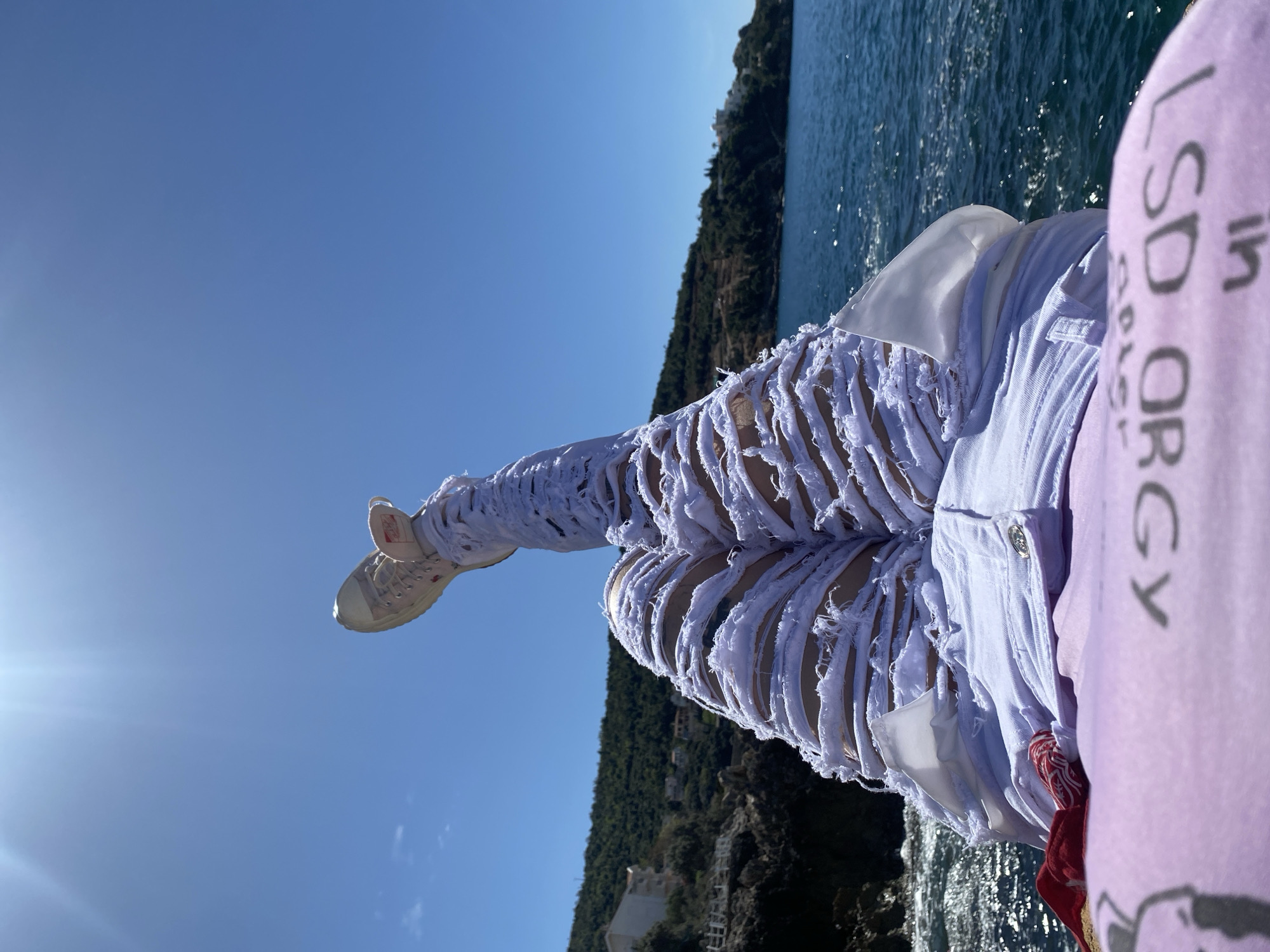 A medium shot captures a person sitting on a rock overlooking the water in Bar, Montenegro. The lower half of the person's body is visible, with legs extended and slightly raised. They are wearing heavily distressed white jeans with numerous rips and tears, along with a pair of pale-colored sneakers. To the right of the image, a portion of a lavender t-shirt is visible. In the background, there is a view of the sea and a treed hill in the distance. The sky is clear and blue.