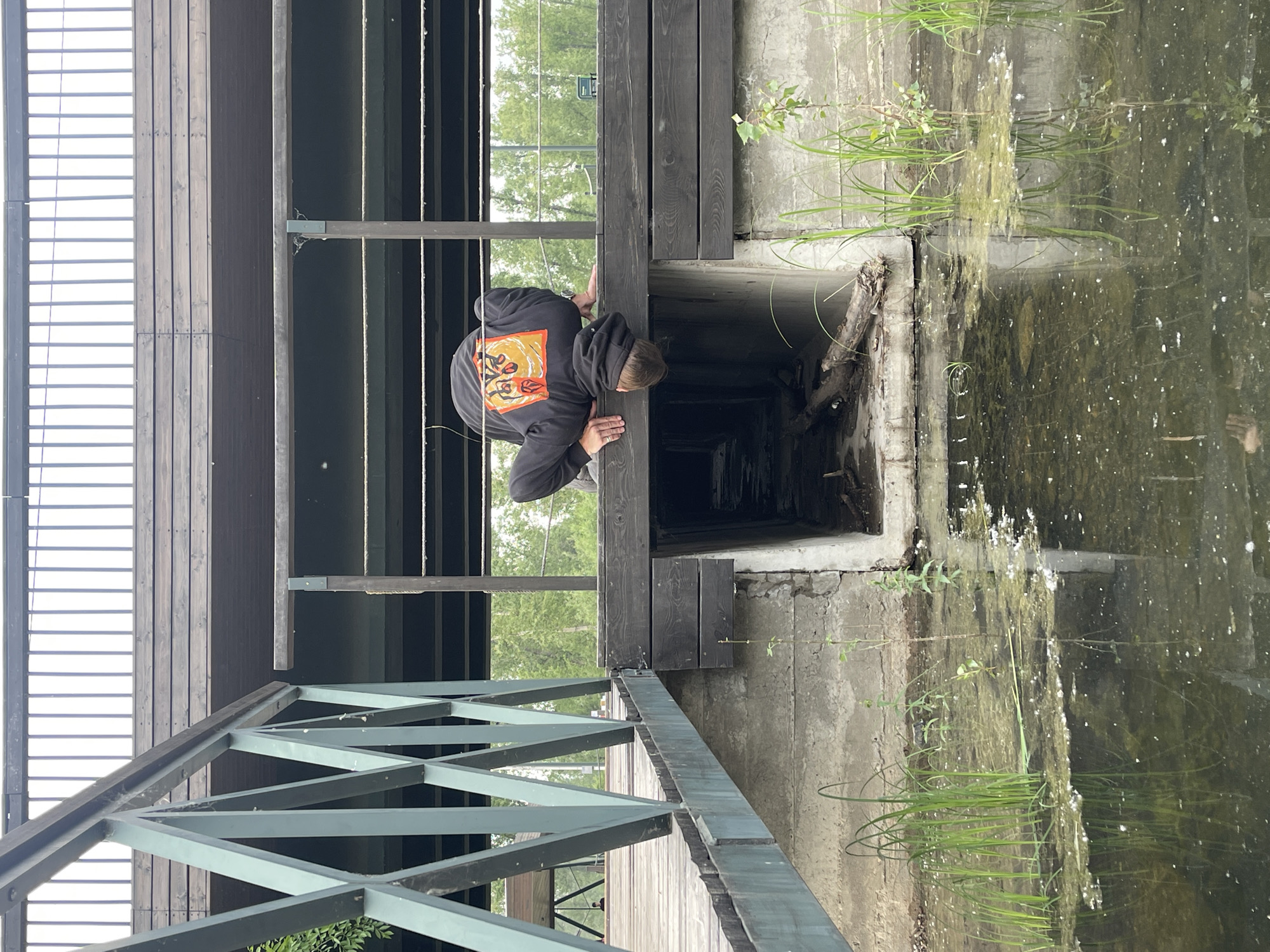 The image shows a person crouching at the edge of a wooden structure, looking down into a dark, rectangular opening. The person is wearing a black hoodie with an orange and yellow graphic design on the back. The structure appears to be part of a bridge or walkway made of dark wood and metal. To the right of the structure is a concrete wall and a body of murky water with green vegetation growing in it. The water reflects the surrounding greenery and the sky. The scene is outdoors, with trees visible in the background. The lighting suggests it is daytime, possibly overcast, as there are no strong shadows. The overall atmosphere is somewhat mysterious and exploratory, given the person's posture and the dark opening they are peering into. The location is identified as Krasnoyarsk, Russia. No visible text is present in the image.