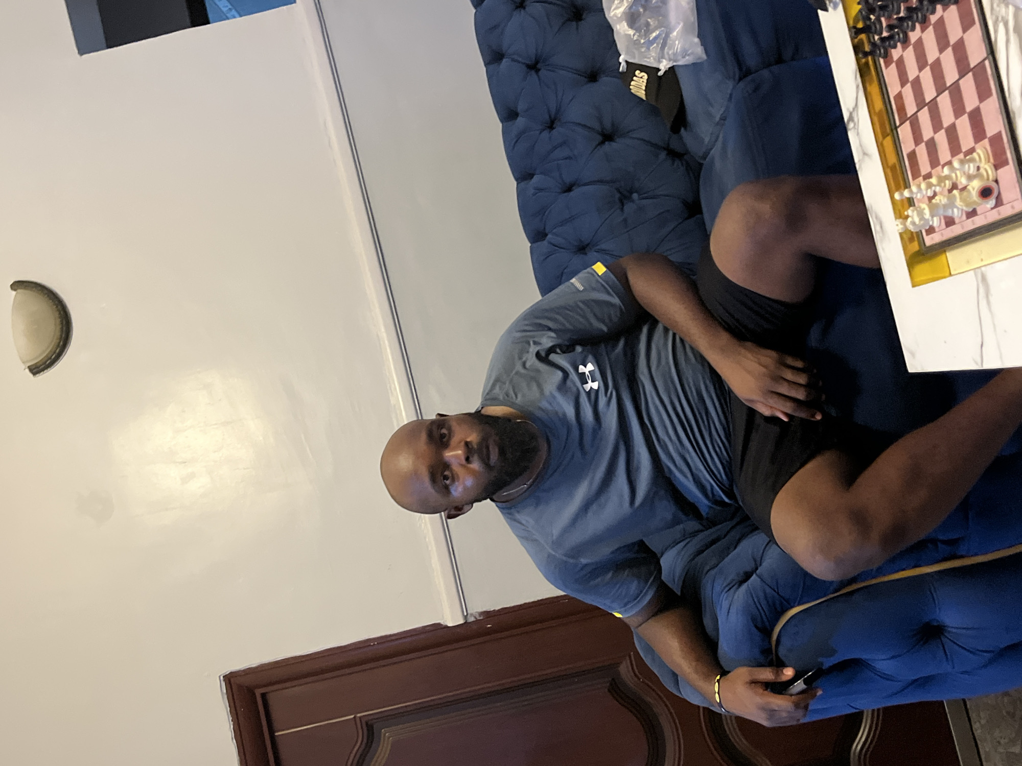 A man is seated on a blue button-tufted sofa. He is wearing a grey Under Armour t-shirt and black shorts. A chess board sits on a table next to him. The background features a light-colored wall, a light fixture, and a dark wood-paneled door. It is not possible to confirm the location as Dushepe, Nigeria from this image.