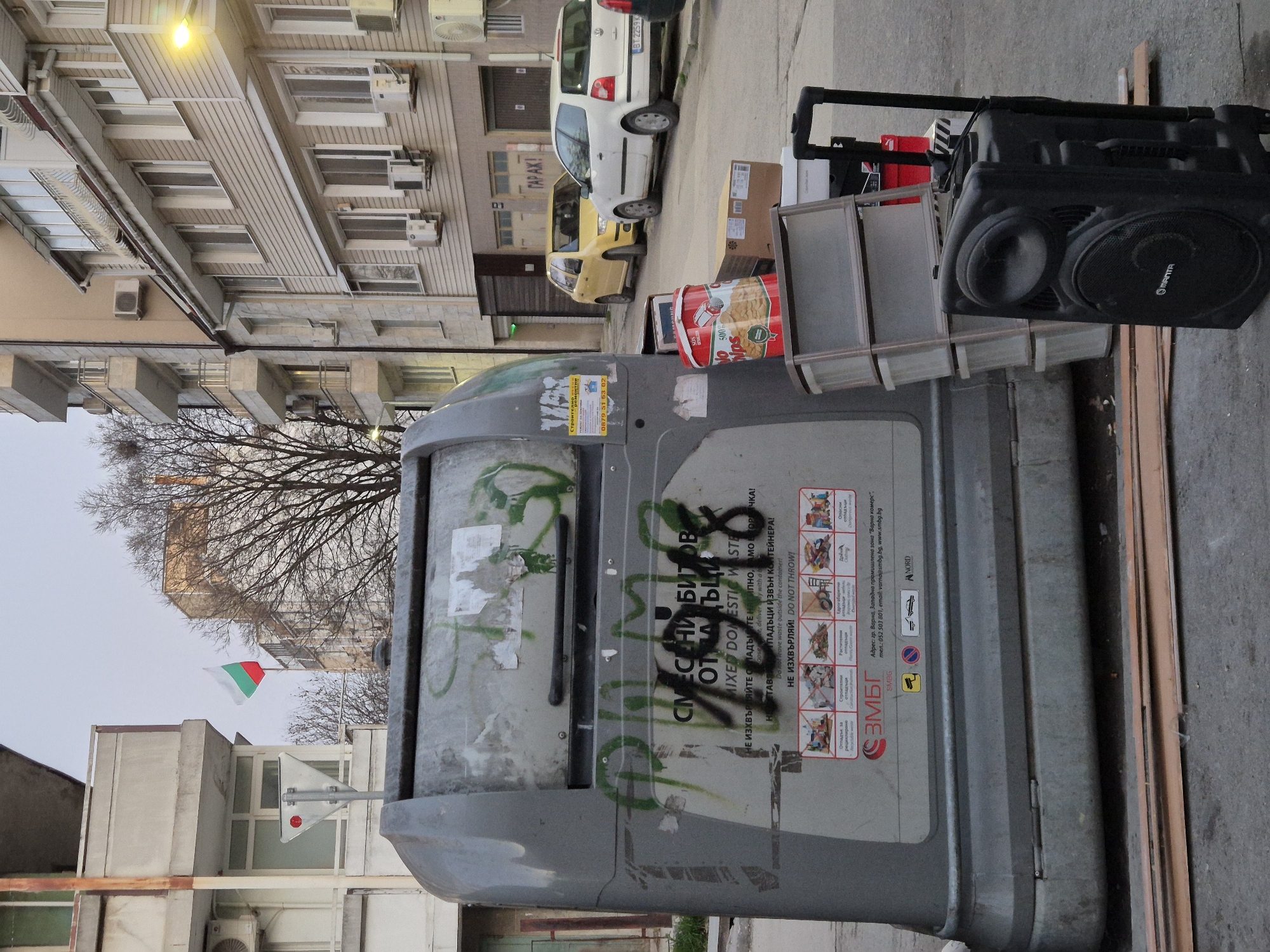 The image shows a street scene in Varna, Bulgaria. A large, gray trash container is in the foreground, covered in graffiti and stickers. Next to the trash bin is a black PA speaker with a carrying handle, and a metal window frame leaning against it. A Bulgarian flag is visible in the background, attached to a pole on top of a building. A leafless tree is visible between the flag and trash container, with buildings on either side. Cars are parked on the street, a white one and a yellow one. The trash container has Bulgarian writing on it, as well as symbols prohibiting certain types of waste.