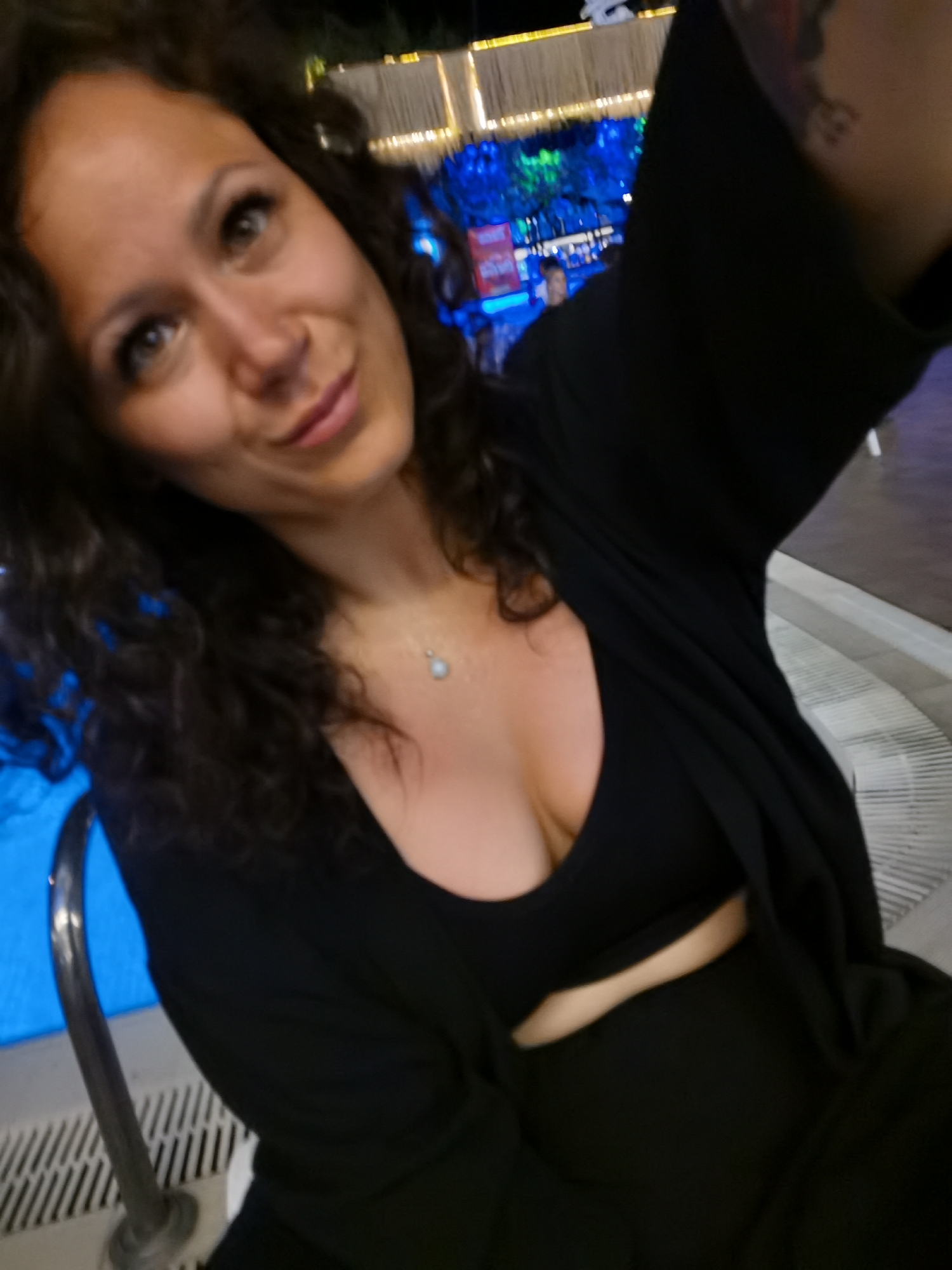 A woman is captured in a selfie-style shot, her face angled towards the camera. She has long, dark, curly hair and her makeup includes defined eyebrows and lashes. She is wearing a black top with a low neckline and a delicate pendant necklace. Her attire also includes a black garment draped over her shoulders. The background suggests an outdoor setting at night, with some illuminated structures and blue lighting visible, indicative of a possibly lively area like a beach bar or entertainment venue in Alanya, Turkey. The overall scene is dimly lit, with the woman's face being the primary focus.