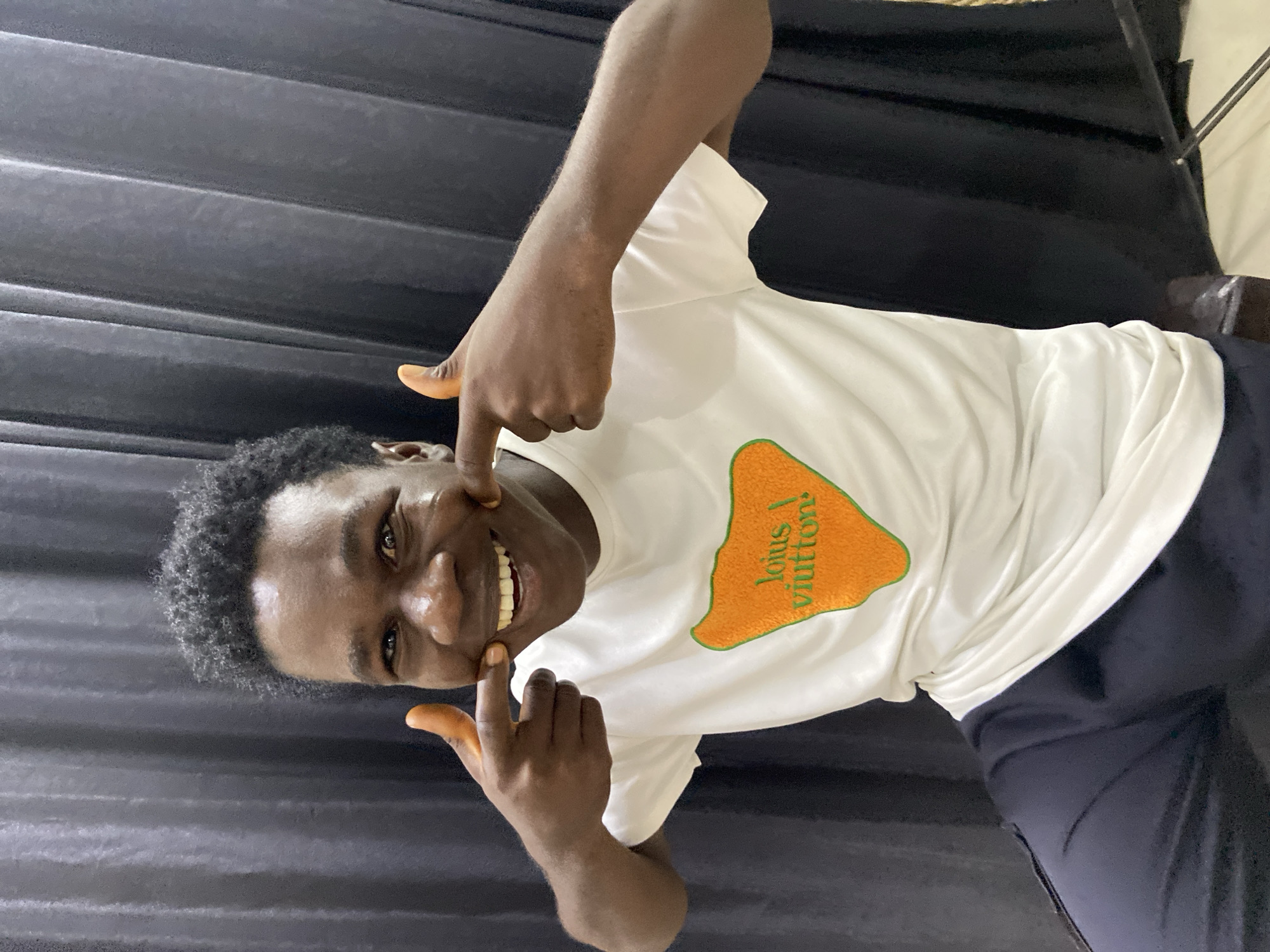 The image displays a young adult male, captured in Mararaba, Nigeria, against a dark grey, vertically pleated curtain backdrop. The individual, exhibiting dark skin and short, dark curly hair, is positioned from the waist up, directly facing the viewer with a wide, toothy smile. Both index fingers are pressed into his cheeks, slightly accentuating his expression. He is dressed in a white, short-sleeved t-shirt featuring an irregular orange patch on the chest, which contains green embroidered text reading 'lolus \ viutton!'. Dark-colored trousers are visible from the waist down. The lower right corner of the frame reveals a partial view of a light-colored surface and a slender dark object, consistent with a stand.