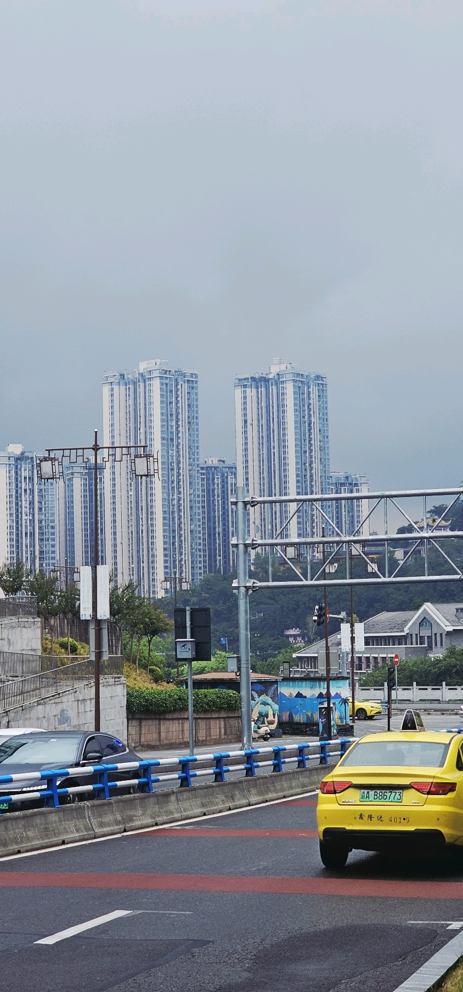 The image captures a street scene in Shapingba District, China, under an overcast sky. In the foreground, a yellow taxi with license plate "A B88773" is driving on a wet asphalt road marked with red and white lines. To the left of the taxi, a dark-colored car is visible behind a concrete barrier with blue and white railings. Further back, a white and blue striped barrier separates the roadway from a sloped walkway with metal railings and some greenery. Several streetlights with rectangular lamps are mounted on poles. In the background, a cluster of tall, modern apartment buildings with numerous windows dominates the skyline. The buildings are a light color with blue accents. Behind the apartment buildings, a lush green, hilly landscape is visible, with some smaller structures scattered on the slopes. A motorcycle is parked to the left of a mural on a small building.