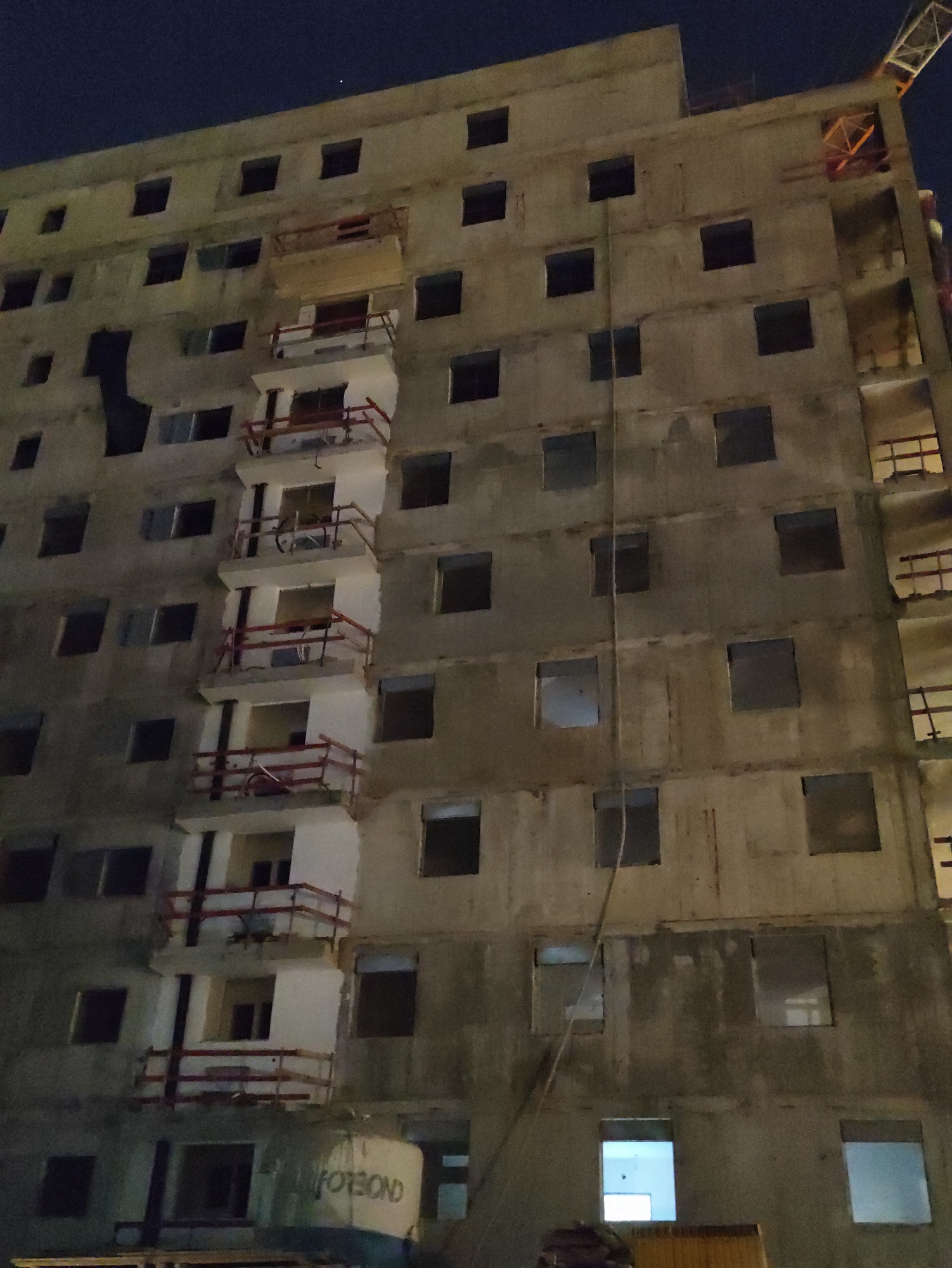 The image shows a multi-story unfinished concrete building at night in Petah Tikva, Israel. The building has multiple rectangular window openings and some balconies with red railings. There is a construction crane visible at the top right of the frame. A rope or cable hangs down the side of the building, and the words "FOTBOND" are visible on a surface near the bottom. The sky above is dark, indicating nighttime. There are no visible people in the image.