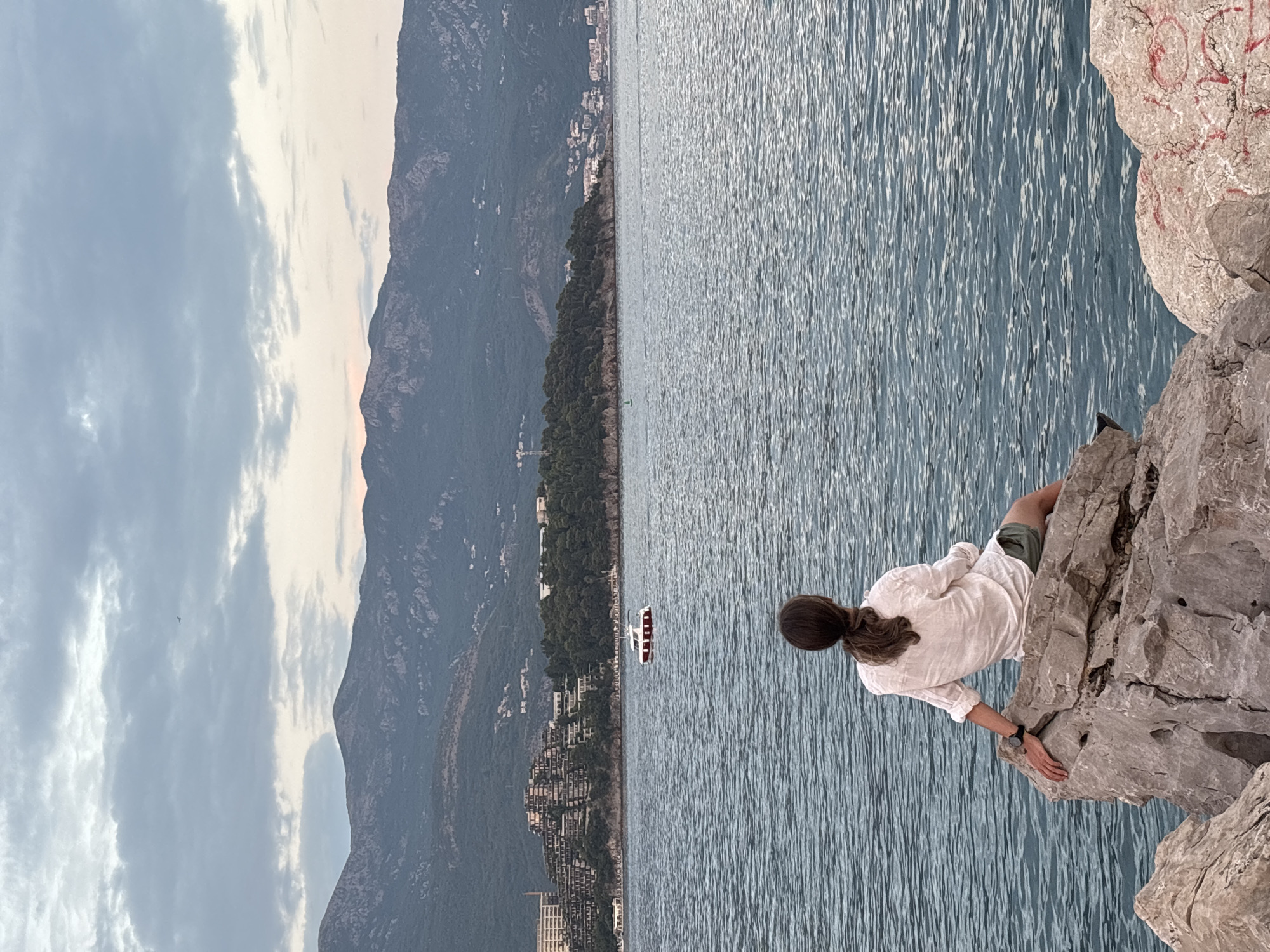 A person with dark hair pulled back in a ponytail sits on large grey rocks overlooking a calm, blue sea. They are wearing a white linen shirt and green shorts. Their left arm rests on the rock, and they wear a black watch on their left wrist. In the distance, a red and white boat travels across the water. Beyond the sea, a densely forested hill is covered with buildings, some of which are terraced into the hillside. Majestic mountains rise in the background under a cloudy sky. The scene appears to be in Budva, Montenegro, likely near the coast with views of the town and surrounding natural landscape.
