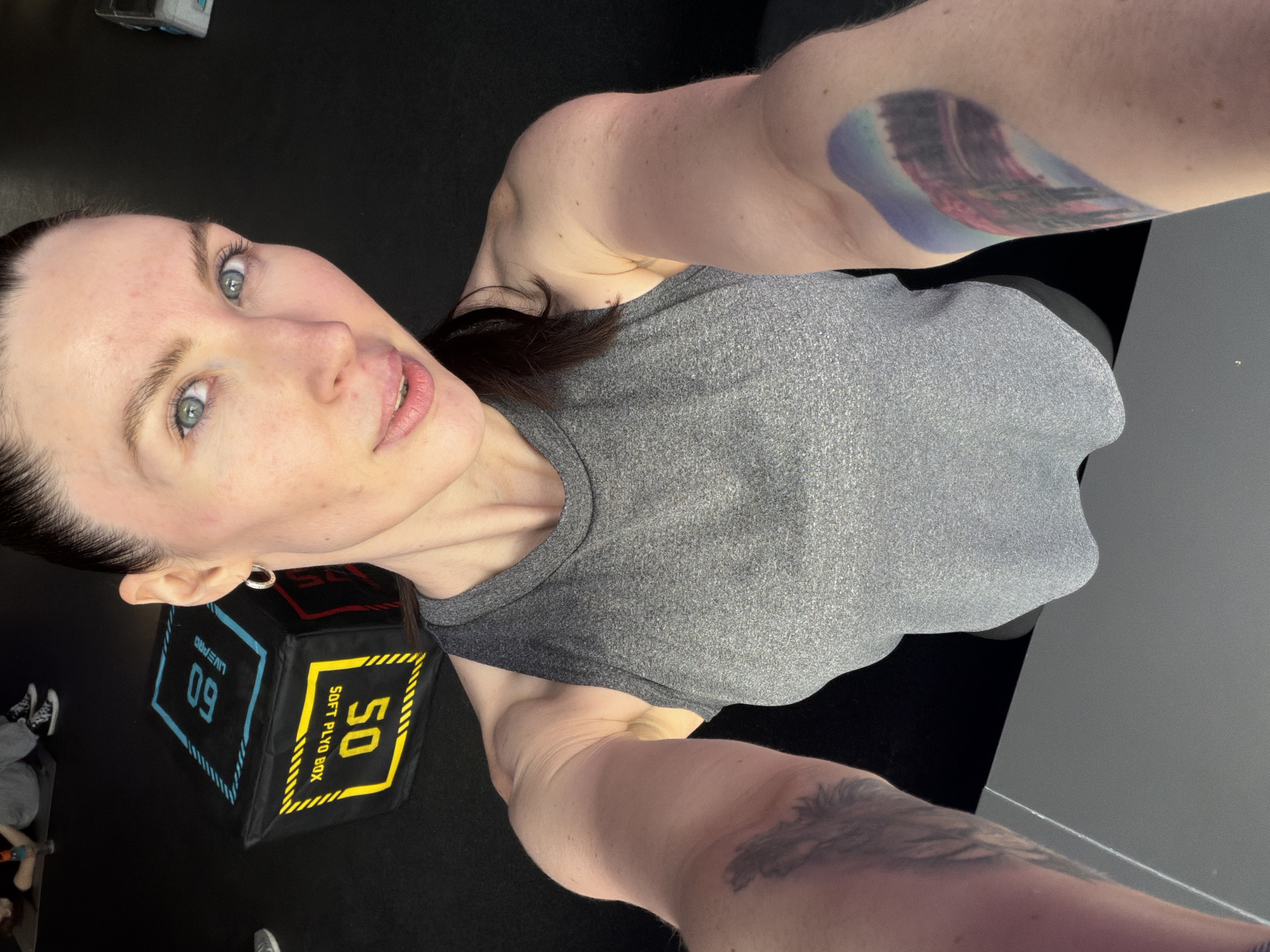 A portrait-oriented selfie captures a woman in an indoor gym setting. The woman is positioned with her head on the left, looking directly at the camera with her body extending towards the right. She has fair skin, light blue-grey eyes, and dark hair pulled back. She wears a grey sleeveless athletic top. Her expression is neutral, and she appears to be engaged with the camera.
Visible on her arms are tattoos; a colorful, possibly abstract or landscape-themed tattoo is on her left upper arm/shoulder, and a darker design is on her right forearm. The background features a dark, likely black, floor and wall, characteristic of a fitness studio. To her left, a black and yellow soft plyometric box is prominently displayed. Text on the box includes "SOFT PLYO BOX" in yellow lettering, along with the numbers "50" (yellow) and "60" (blue), and the brand "LIVE PRO" in blue. Hints of other people's feet or legs are visible in the background near the plyo box. The scene is illuminated by artificial indoor lighting. The event taking place is a self-portrait session within a gym, located in Perm, Russia.