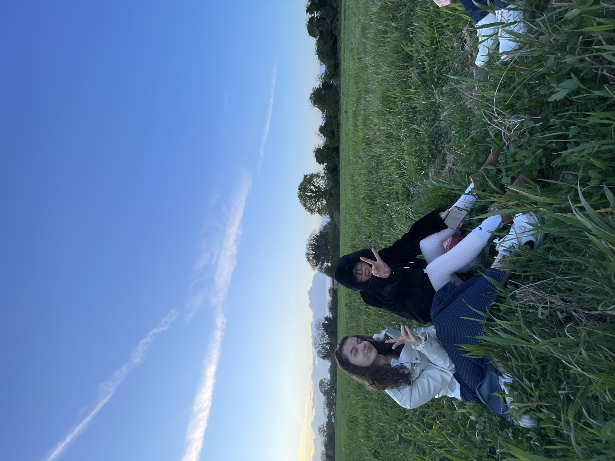 Two young women are lying in a grassy field. The woman on the bottom wears a light-colored jacket and dark pants, while the woman on top wears a black hooded sweatshirt and light pants. Both are making peace signs with their fingers. The sky is blue with a few clouds, and there are trees in the background. The location is Maidstone, United Kingdom.