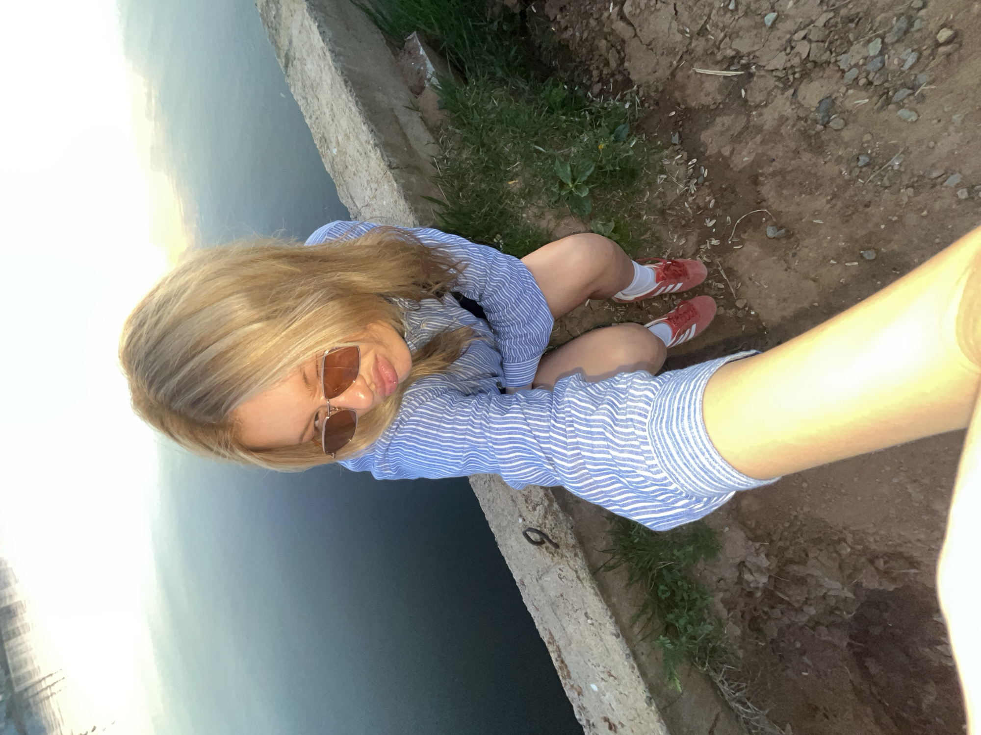 A blonde woman wearing a blue and white striped shirt, sunglasses, and red shoes is sitting on the edge of a concrete structure. Water is visible behind her, and dirt with some grass is beside her. The photograph appears to be a selfie, with the photographer's arm visible in the lower right corner. It is unknown if the photograph was taken in the city of Chaikovsky, Russia.