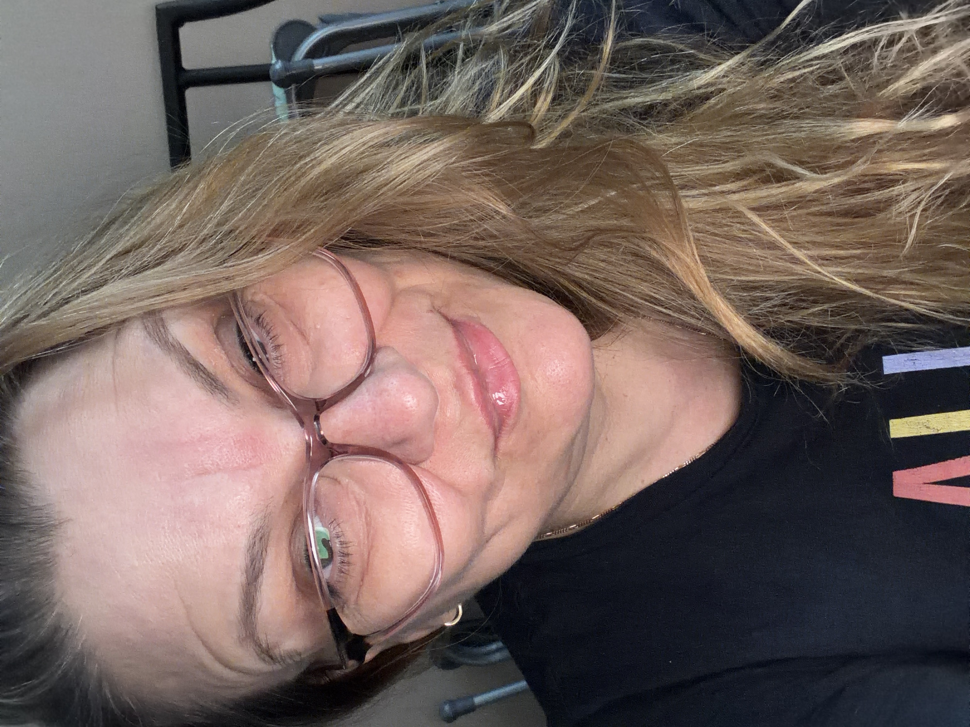 This is a close-up, eye-level, self-portrait of a Caucasian woman with blonde hair wearing pink-rimmed glasses and a black t-shirt. She is looking slightly off-camera to her right with a subtle smile. The lighting is warm and highlights her facial features, including fine lines on her forehead and around her eyes. Her hair is styled loosely around her face and cascades over her shoulder. A subtle gold chain is visible around her neck. In the background, out of focus, are what appear to be metal structures, possibly furniture or gym equipment, and a dark frame. The scene is indoors, with no discernible features that would place it specifically in Southend-on-Sea, United Kingdom.
