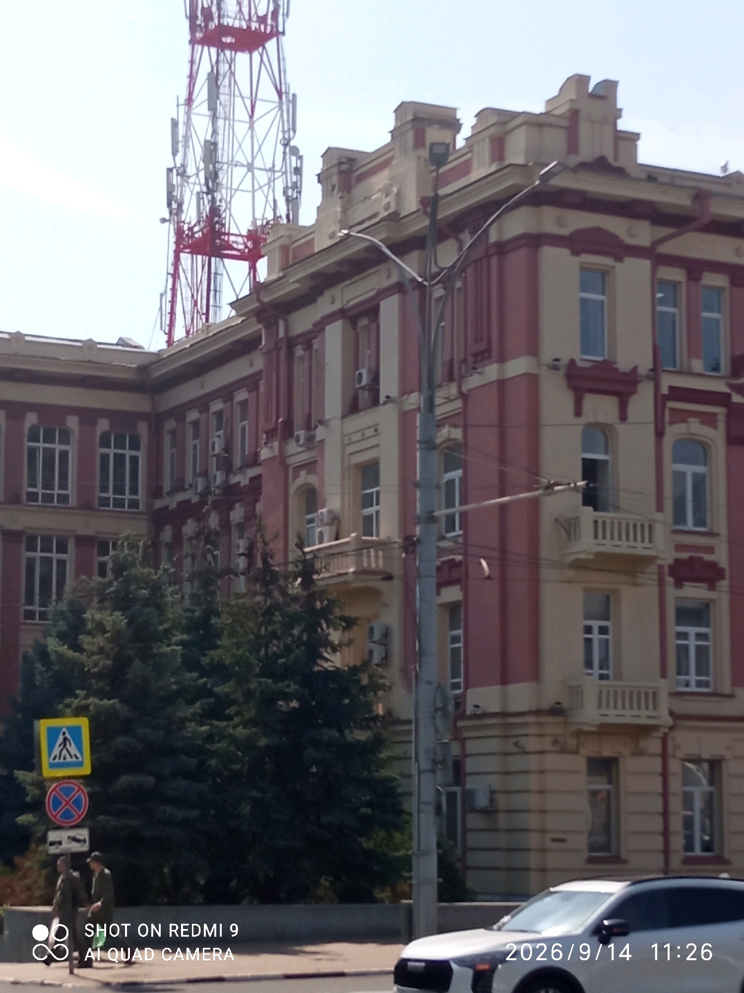 The image shows a multi-story building with a red and cream facade, featuring architectural details such as decorative cornices and balconies. A large communication tower with red and white lattice work stands behind and above the building. In the foreground, there are traffic signs indicating a pedestrian crossing and a prohibition, along with two individuals walking on the sidewalk. A white car is visible on the road. The scene appears to be in an urban environment, with trees lining the sidewalk. The timestamp on the image indicates September 14, 2026, at 11:26 AM. The watermark "SHOT ON REDMI 9 AI QUAD CAMERA" suggests the photo was taken with a Redmi 9 smartphone. Based on the architectural style of the building, the presence of the communication tower, and the overall urban setting, this scene is likely located in Rostov-on-Don, Russia.