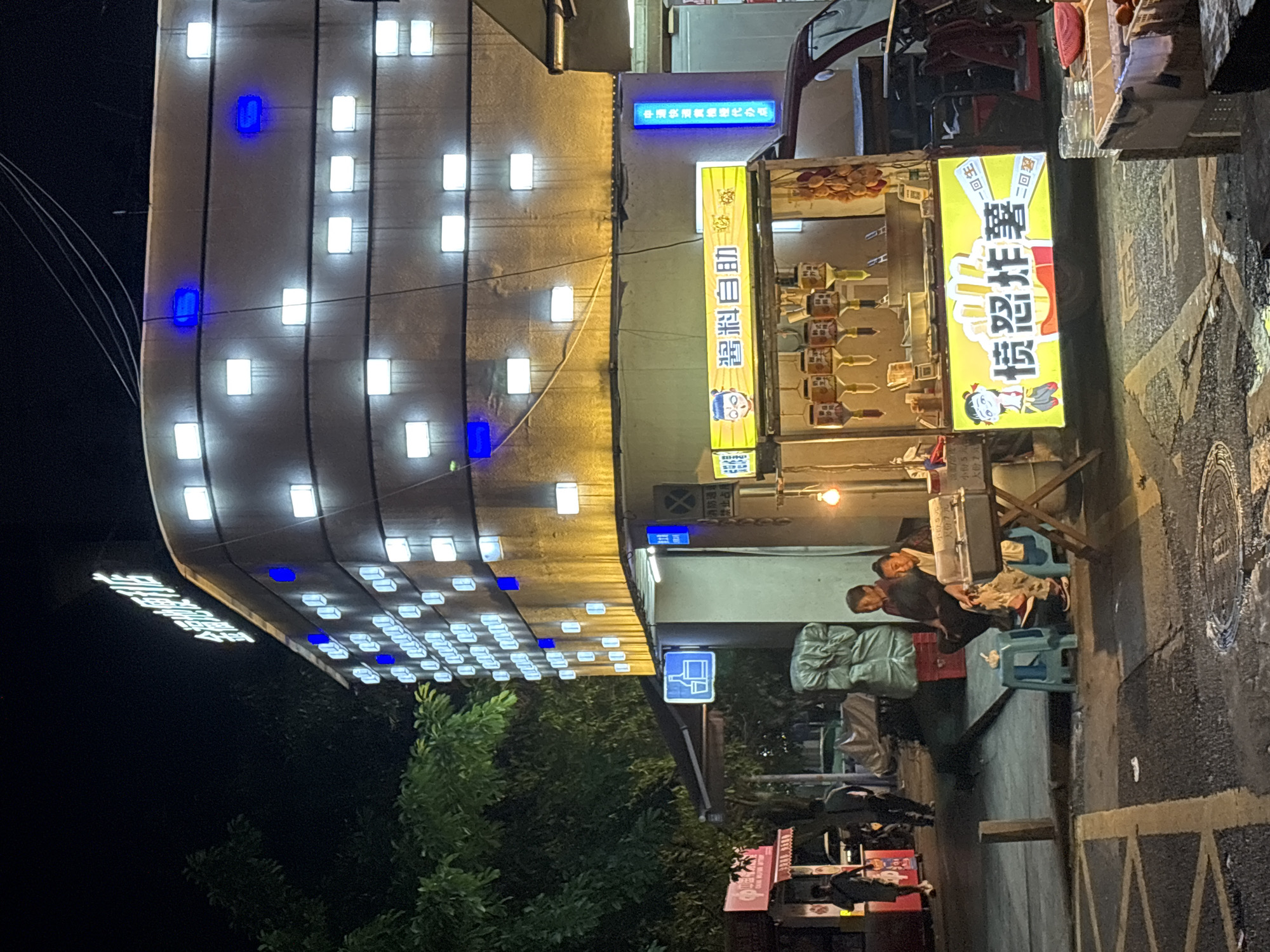 Here is a description of the image.
The image shows a nighttime street scene in Nan'an District, China. A brightly lit building is prominent on the left. It's decorated with numerous square lights, some of which are blue. Several signs are attached to the building, including one for a food stall with Chinese characters "愤怒炸薯", which translates to "Angry Fried Potato". The food stall also has a sign saying "酱料自助", or "Sauce Self-Service". Two people are sitting nearby, possibly customers of the food stall. A traffic or directional sign is partially visible below the building. A small store or vending machine is located in the lower part of the image. Street details, including the curb and pavement, are also visible.