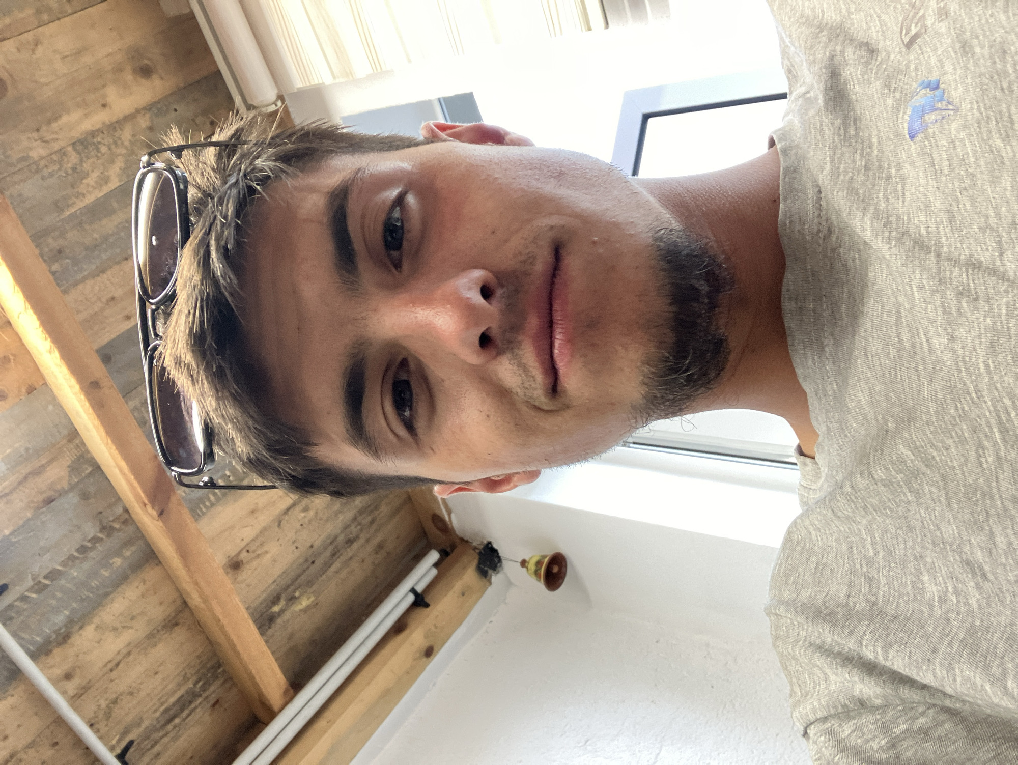 A selfie shot shows a man with short brown hair, glasses perched on his head, and a light stubble beard. He is wearing a gray T-shirt. The background includes a white wall with a window, exposed wooden ceiling beams, and white pipes attached to the wooden beams. A small brass bell hangs from the ceiling. No location in the city of Komuniga, Bulgaria, is identifiable.
