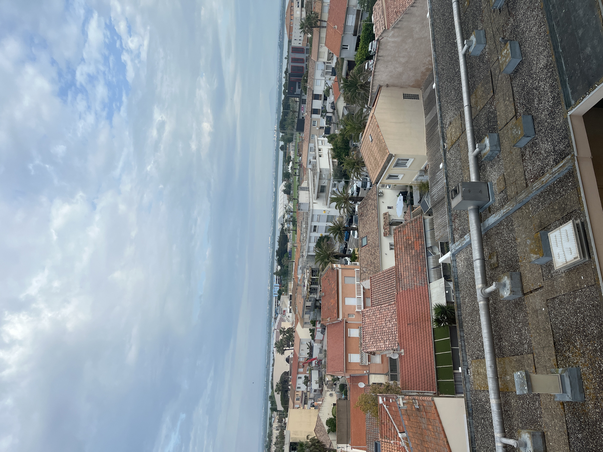 This is an eye-level shot of Palavas-les-Flots, France, taken from a high vantage point, likely a rooftop or high floor of a building. The sky is partly cloudy with a mix of blue sky and grey clouds. Below, the Mediterranean Sea is visible as a flat, grey plane stretching towards the horizon.
The city of Palavas-les-Flots consists of a densely packed collection of buildings, mostly two-to-three stories tall, with a few taller structures. The buildings are a mix of white and peach-colored walls with predominantly red tile roofs. Green trees and palm trees are interspersed throughout the cityscape.
The foreground is dominated by the flat gravel rooftop. Grey metal piping runs vertically along the roof. Square concrete blocks are positioned at intervals, likely used for support or securing the piping. There are also some additional vent structures on the roof.