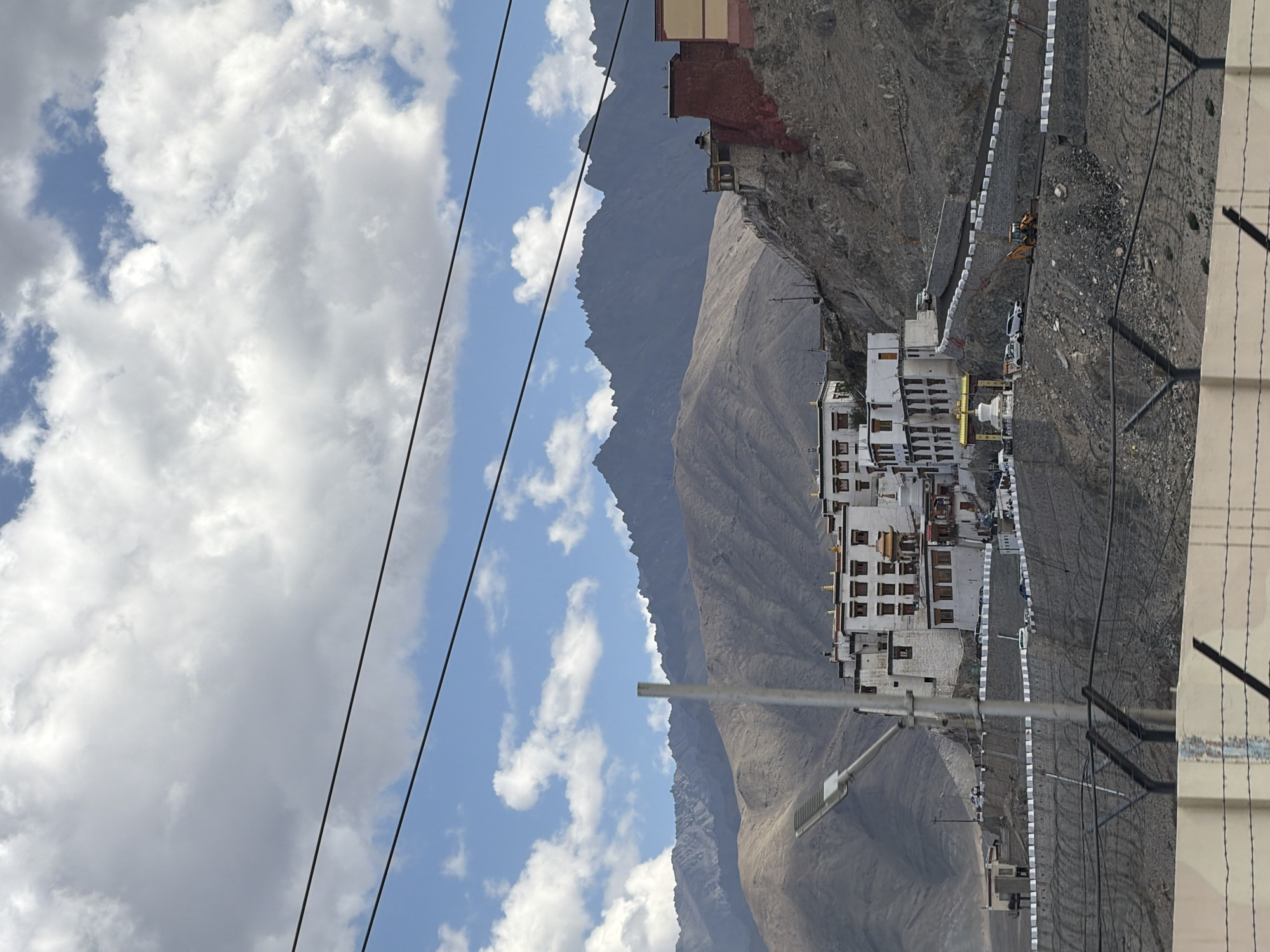 The image shows a panoramic view of a monastery perched on a hillside in Spituk, India, with a mountainous backdrop and a cloudy sky. The monastery complex consists of several white buildings with red-trimmed windows and roofs, built in a traditional architectural style. A golden stupa and an ornate entrance gate are visible in the foreground. The hillside is covered with rocky terrain and sparse vegetation. A paved road winds its way up the hill, with several vehicles parked along it. A yellow excavator is seen working on the road. Power lines crisscross the sky in the upper portion of the image. The foreground is partially obscured by a barbed wire fence.