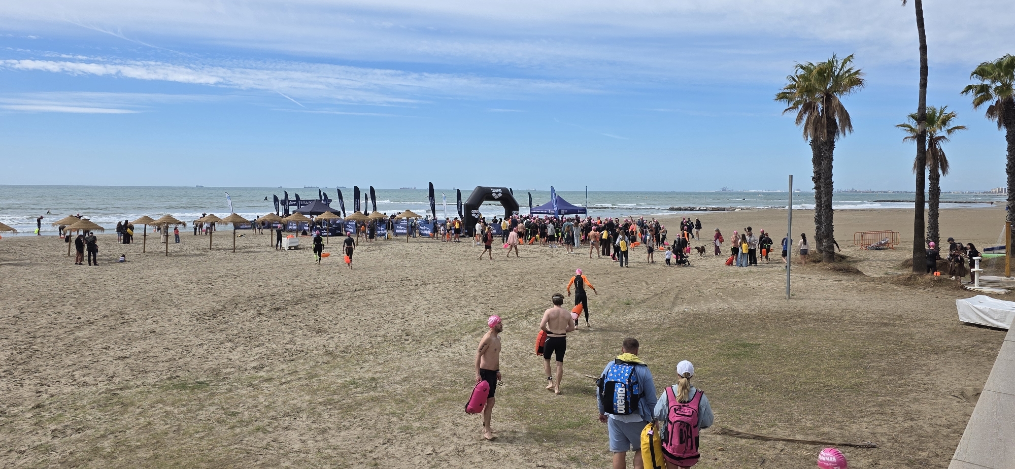 This image depicts a beach scene in Benicàssim, Spain. The sky is blue with scattered clouds, and the sea is visible in the background with several ships. A sandy beach stretches across the foreground and midground.
There is a gathering of people near a black inflatable arch labeled "Arena," presumably an event setup for a swimming or triathlon event. Some people are wearing pink swim caps and carrying safety floats, indicating they are participants. There are tents, flags, and umbrellas scattered around the event area.
In the foreground, several individuals are walking away from the event area. Some are shirtless men carrying safety floats; others wear backpacks. Palm trees line the right side of the image, and a building is partially visible in the bottom right corner. Overall, the scene appears to be a sunny day at the beach with an event taking place.