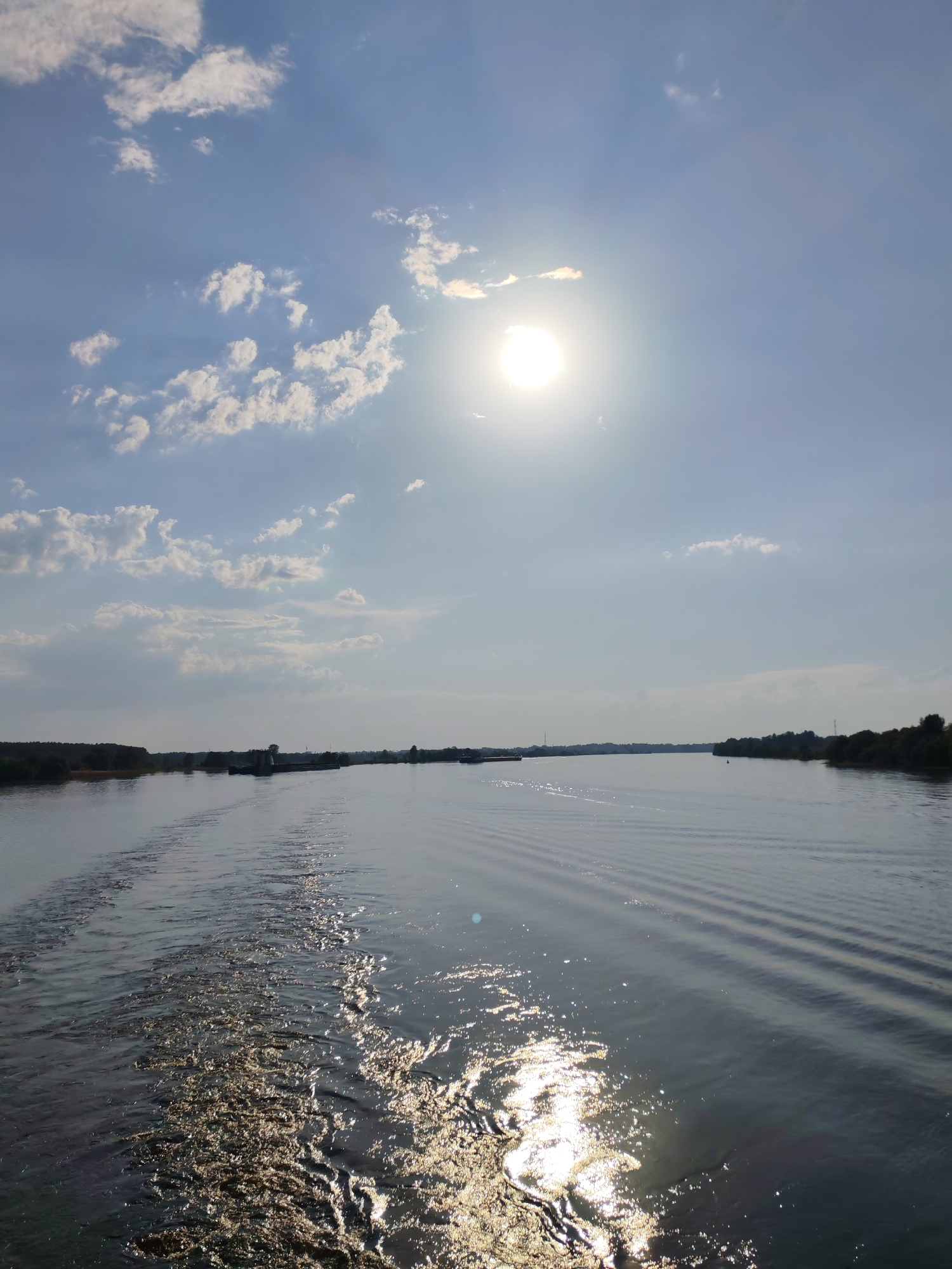 The image depicts a river scene in Titovo, Russia, under a bright, sunlit sky with scattered clouds. The sun is positioned high in the sky, casting a strong glare. The river surface is reflective, displaying patterns of ripples and waves, with concentrated reflections of sunlight. The riverbanks are lined with trees and vegetation, with what appears to be a barge or large vessel visible in the distance, as well as a transmission tower. The viewpoint is likely from a moving boat, evidenced by the wake in the foreground.