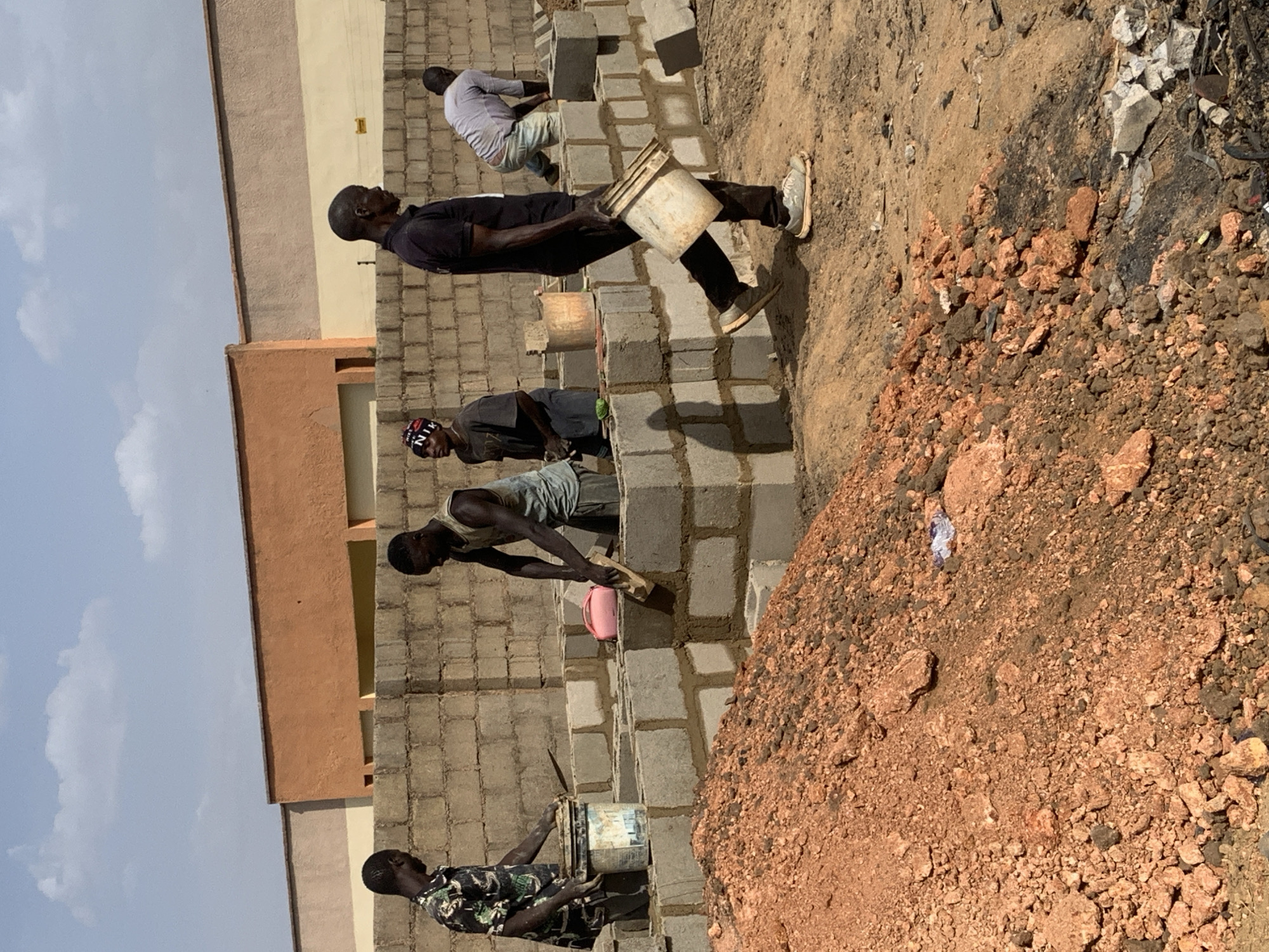 In Rafin Bauna, Nigeria, a construction site shows four men laying concrete blocks to build a wall. They are working outdoors, near a pile of red dirt and rubble. The building under construction is beige and orange. One man carries a bucket, another uses a trowel, and others appear to be moving materials and setting blocks. The sky is visible with scattered clouds.
