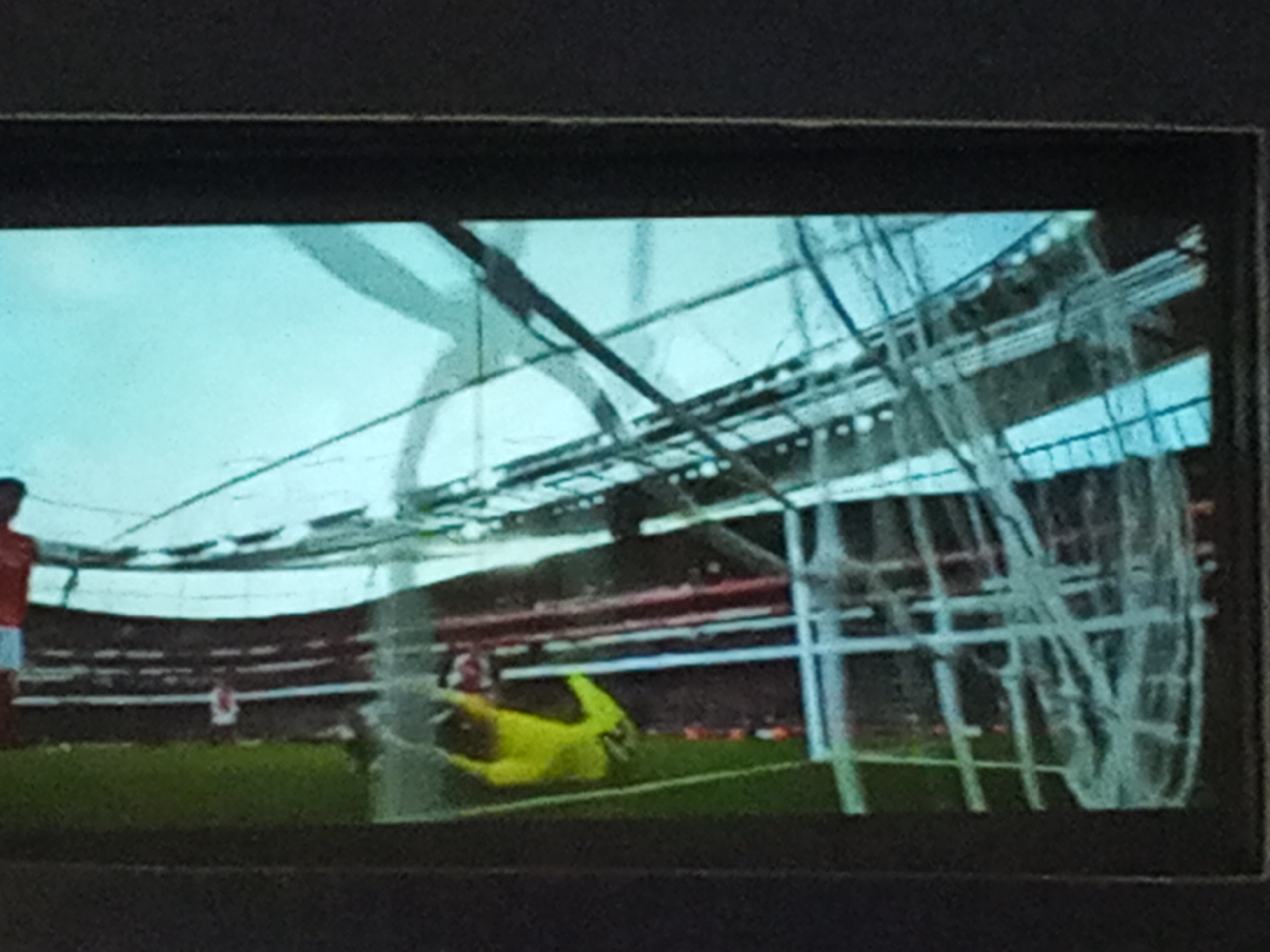 The image shows a soccer game in progress, viewed through the netting of a goal. A goalkeeper in yellow is diving to make a save. Other players in red and white are on the field. The game is taking place in a stadium with red seats. The city and country of the location cannot be determined from the image.