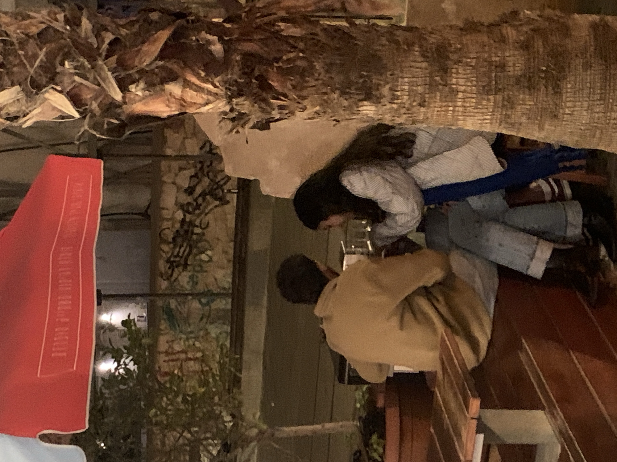 Two people are seated at a wooden table outdoors. The woman has long dark hair and is wearing a light-colored striped shirt and blue jeans. The man is wearing a tan jacket. They appear to be engaged in an activity, possibly reading or working. The scene is lit by artificial lighting. A palm tree trunk is overhead, with dried fronds extending into the frame. In the background, there is a building with an exterior wall made of stone. A red advertisement or sign is partially visible. Based on the style of the building and vegetation, the location appears to be in Tel-Aviv, Israel.