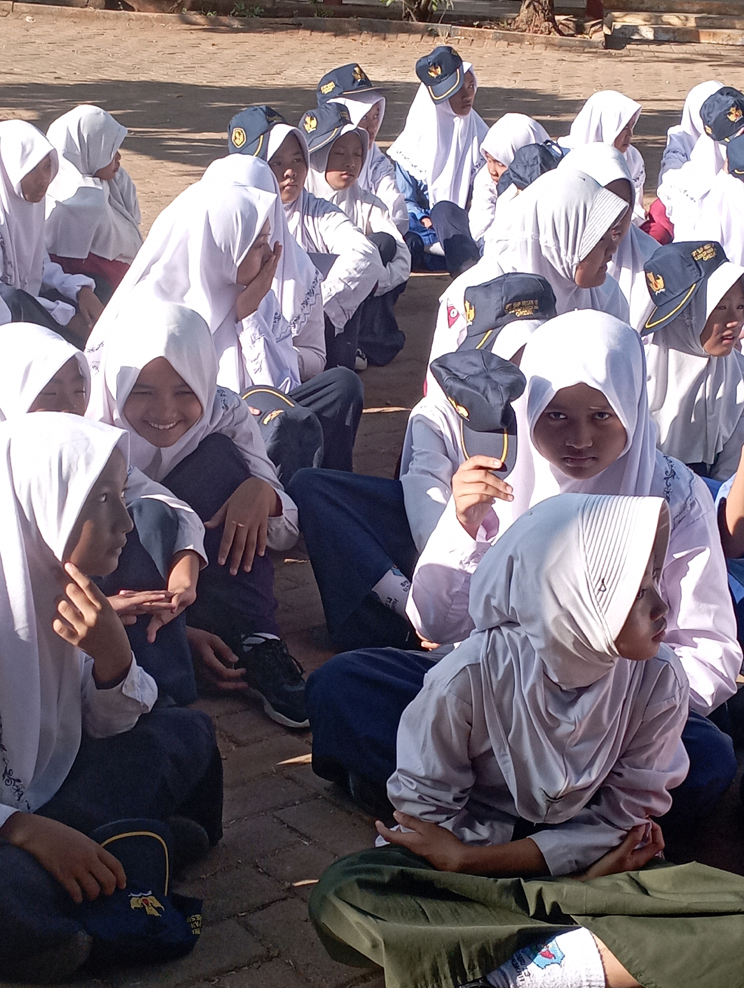 The image depicts a group of young people, likely students, sitting on the ground outdoors. They are all wearing white headscarves and long-sleeved tops, with some also wearing dark-colored pants or skirts. Several individuals are wearing dark caps with yellow trim and emblems, suggesting a uniform or school affiliation. The ground appears to be paved with reddish-brown bricks or tiles, and there is some foliage and a tree visible in the background, indicating an outdoor educational or recreational setting. The lighting suggests it is daytime and sunny, with shadows cast on the ground. The individuals are looking in various directions, and some appear to be interacting with each other, with one person in the foreground smiling and looking towards the camera. The presence of school-related caps and attire suggests this scene may be taking place at a school in Banyuurip, Indonesia.