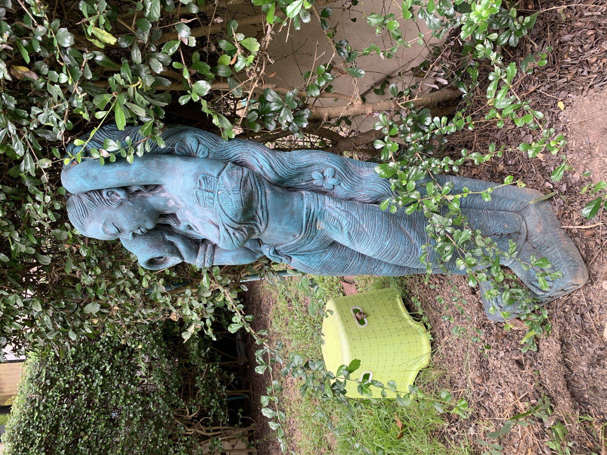 In Baan Bo Phut, Thailand, a weathered, blue-patinaed statue of a woman is partially obscured by lush green foliage. The statue depicts a woman with her arms raised, holding a large jug or pot to her head. She wears a draped garment around her torso and lower body, adorned with intricate patterns. A small, green plastic stool sits to the left of the statue, in the grass. The ground surrounding the statue is covered in mulch and dry leaves, suggesting an outdoor, possibly garden or yard, setting. The dense vegetation and the statue's aged appearance create a sense of time and nature reclaiming the object.