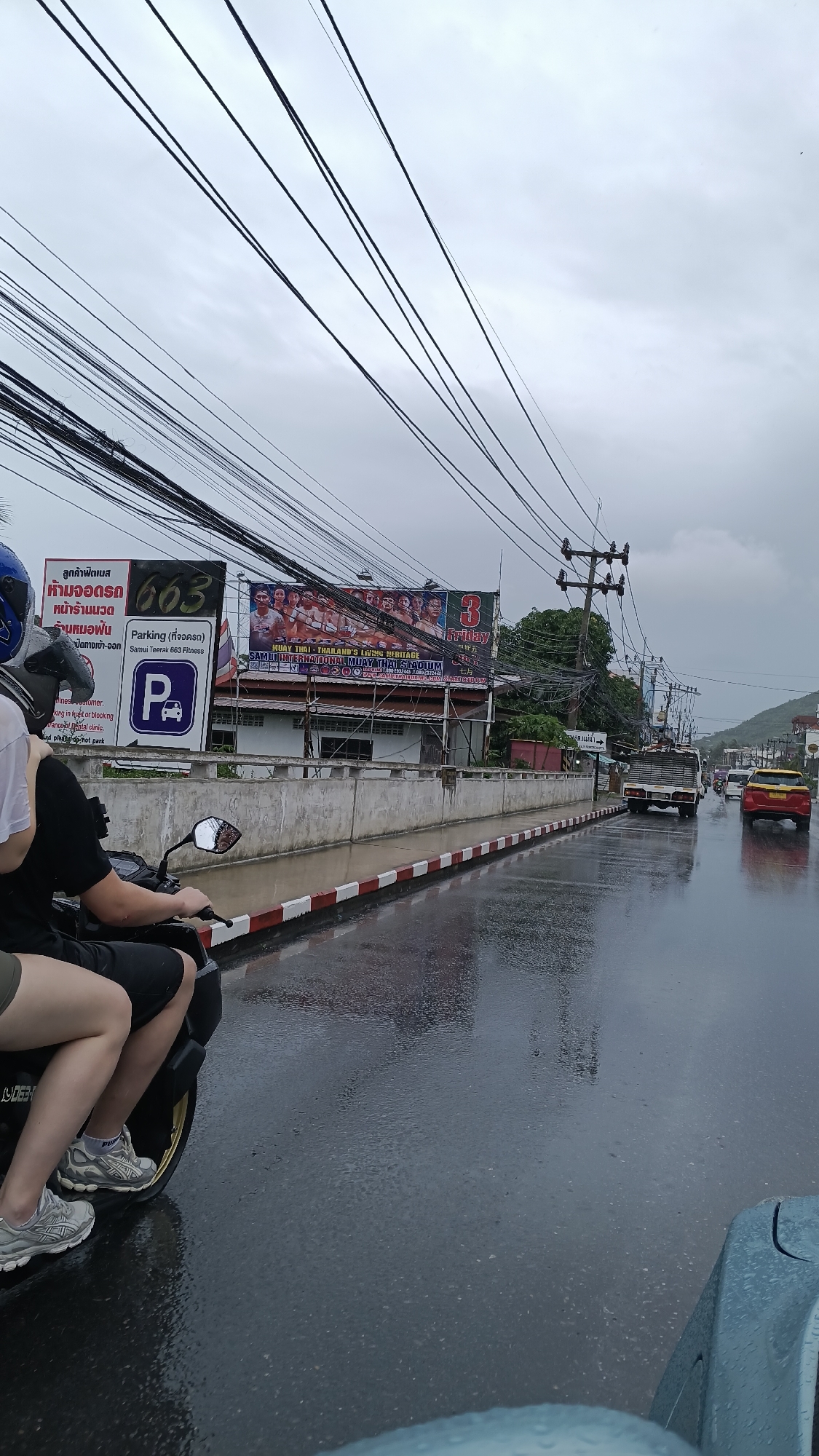 A low-angle, wide shot captures a rainy day on a street in the Ko Samui District, Thailand. The foreground features the rear of a motorcycle with two people riding. The person in front is wearing a black t-shirt and black shorts, while the person behind them is wearing a white t-shirt and khaki shorts. Both are wearing athletic shoes. The motorcycle's front wheel is partially visible. The wet asphalt road reflects the overcast sky and surrounding buildings.
Along the side of the road, a low concrete wall topped with red and white striped barriers separates the sidewalk from the road. A billboard is visible above the wall, displaying advertisements. One billboard features Thai text and the number "663". Another larger billboard shows a group of people and text including "MUAY THAI THAILAND'S LIVING HERITAGE" and "SAMUI INTERNATIOANL MUAY THAI STADIUM".
Further down the street, a white truck with a cargo bed and a yellow sedan are driving on the wet road. Overhead, a dense network of electrical wires crisscrosses the sky, supported by utility poles. In the distance, a hillside with vegetation is visible under the cloudy sky. The overall scene suggests a typical day on a street in Ko Samui, with the rain adding a layer of wetness and reflective surfaces.