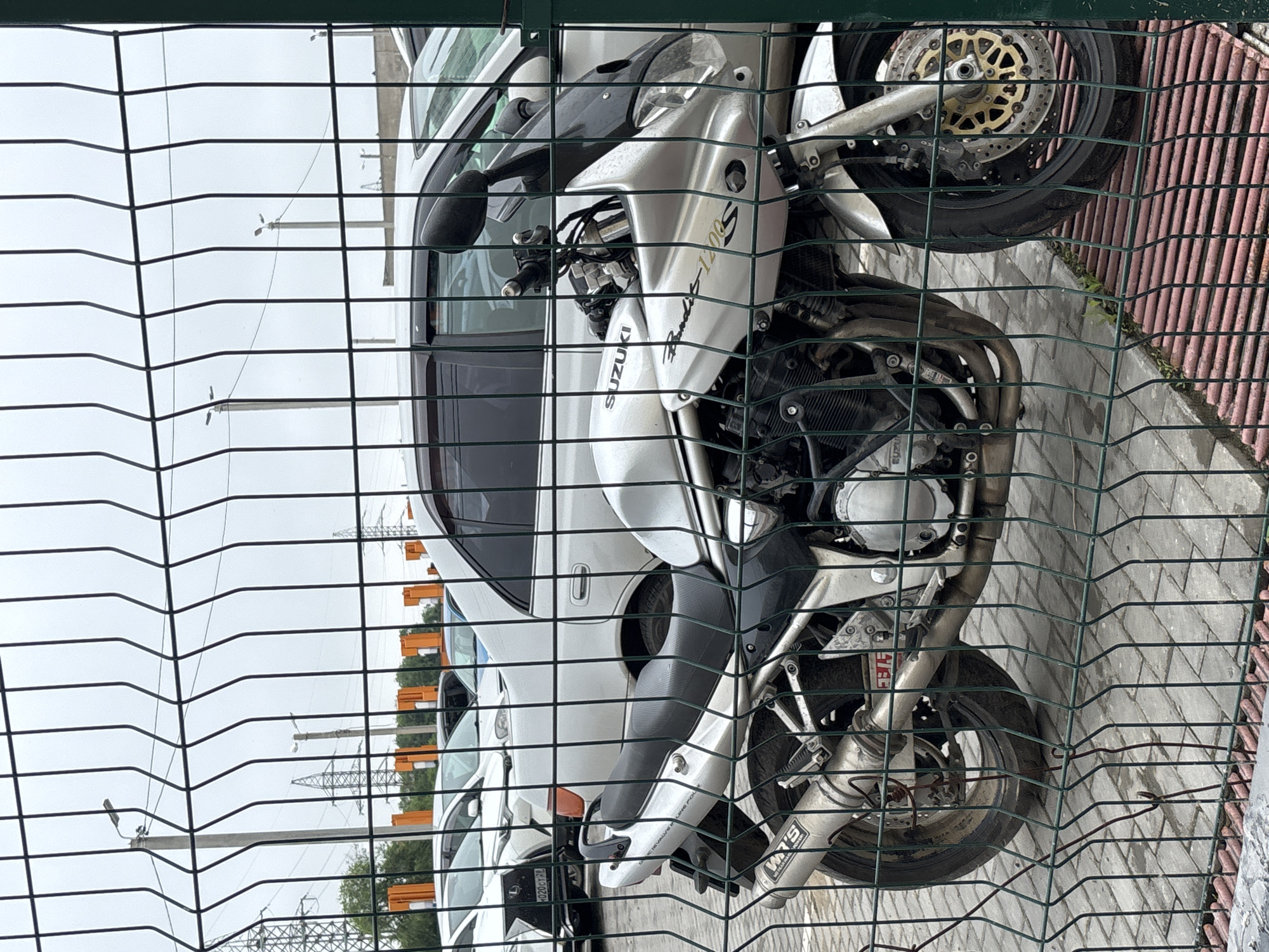 The image shows a silver Suzuki Bandit 1200S motorcycle parked behind a green wire fence. The motorcycle is positioned in the foreground, with its front wheel slightly turned to the right. The seat is black, and the exhaust pipe is silver with a "Yoshimura" logo visible. In the background, there are several cars parked, some of which appear to be white sedans. The ground is paved with grey interlocking bricks, and a red metal grate is visible in the lower right corner. Several utility poles with lights and power lines are visible in the distance, suggesting an urban or suburban environment. There are no people visible in the image. The overall scene appears to be a parking lot, possibly at a dealership or storage facility. Based on the context provided by the user, this scene is located in the city of Blagoveshchensk, Russia.
