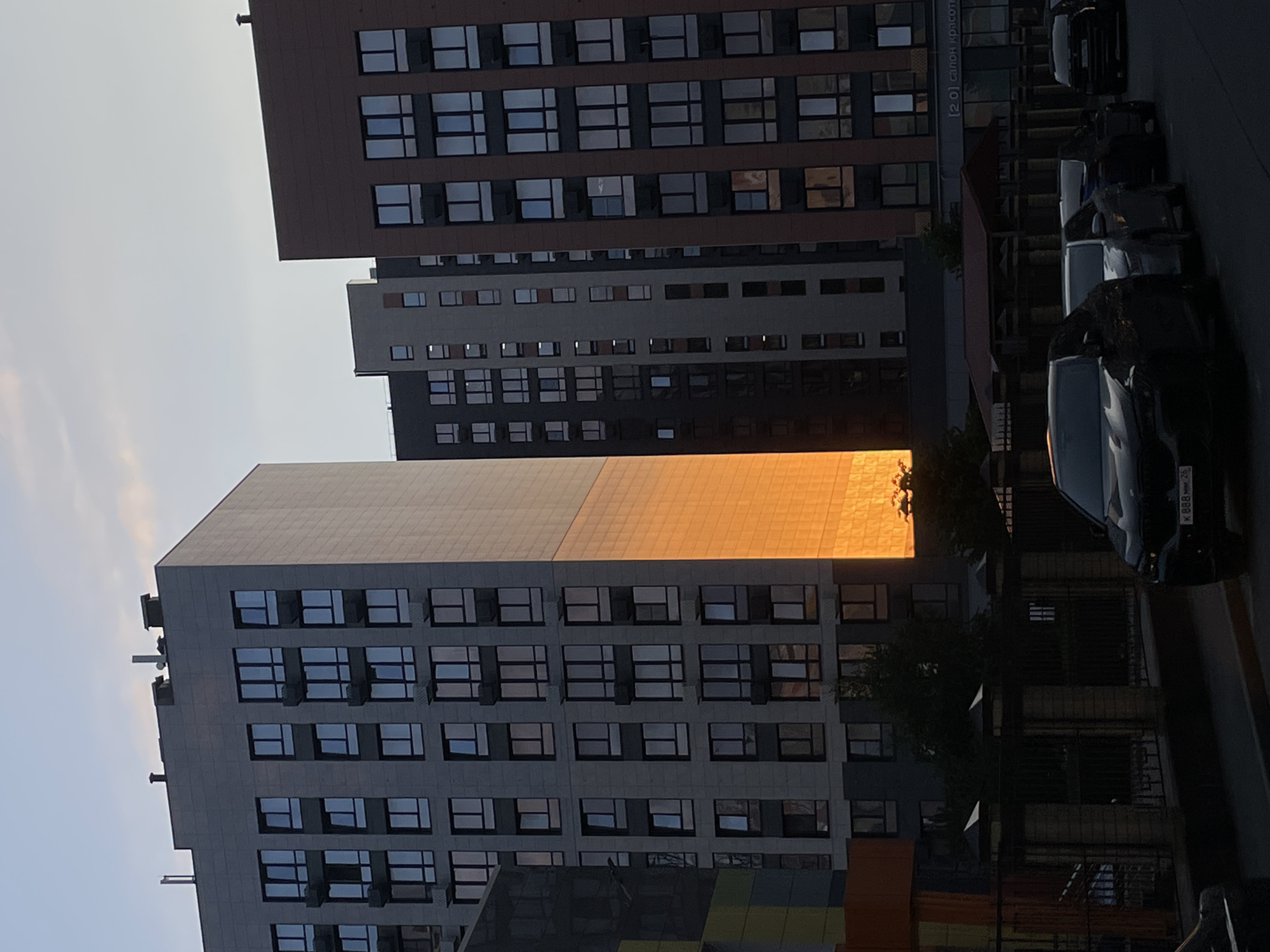 The image depicts a street scene in Stavropol, Russia, with a focus on modern residential buildings. The sky above is a soft gradient of pale blue and pink, suggesting either sunrise or sunset. Several multi-story buildings are visible, characterized by their rectangular shapes and numerous windows. One building on the left features a facade with a distinct section bathed in the warm, golden light of the setting or rising sun, creating a strong visual contrast with the surrounding grey and darker facades.
In the foreground, a dark-colored car with a license plate visible (K 888 MH 26) is parked on the street, along with other vehicles. A fence with brick posts and metal bars runs along the sidewalk, partially obscuring the view of the buildings behind it. Trees are sparsely scattered between the buildings and along the street. The overall impression is one of urban architecture in a tranquil, late-day setting.