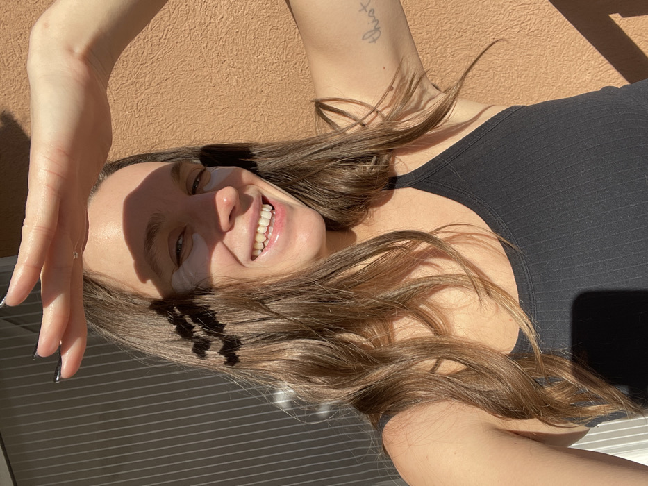 A single female subject is depicted in a brightly lit outdoor environment, likely in Budva, Montenegro. She has medium-length brown hair, a fair complexion, and is smiling broadly, revealing her upper teeth. Two light-colored eye patches are visible under each eye. Her right hand is raised above her forehead, palm forward, positioned to shade her eyes from the sun, casting distinct shadows from her fingers onto her face. The subject is wearing a black, ribbed tank top or sports bra. A faint, light-colored tattoo, appearing to be text, is visible on the inside of her left bicep. The background consists of a textured, light orange or terracotta-colored wall on the right side, and a darker, vertically grooved or ridged surface on the left, possibly a closed blind or architectural detail. Strong sunlight illuminates the scene, creating prominent shadows.