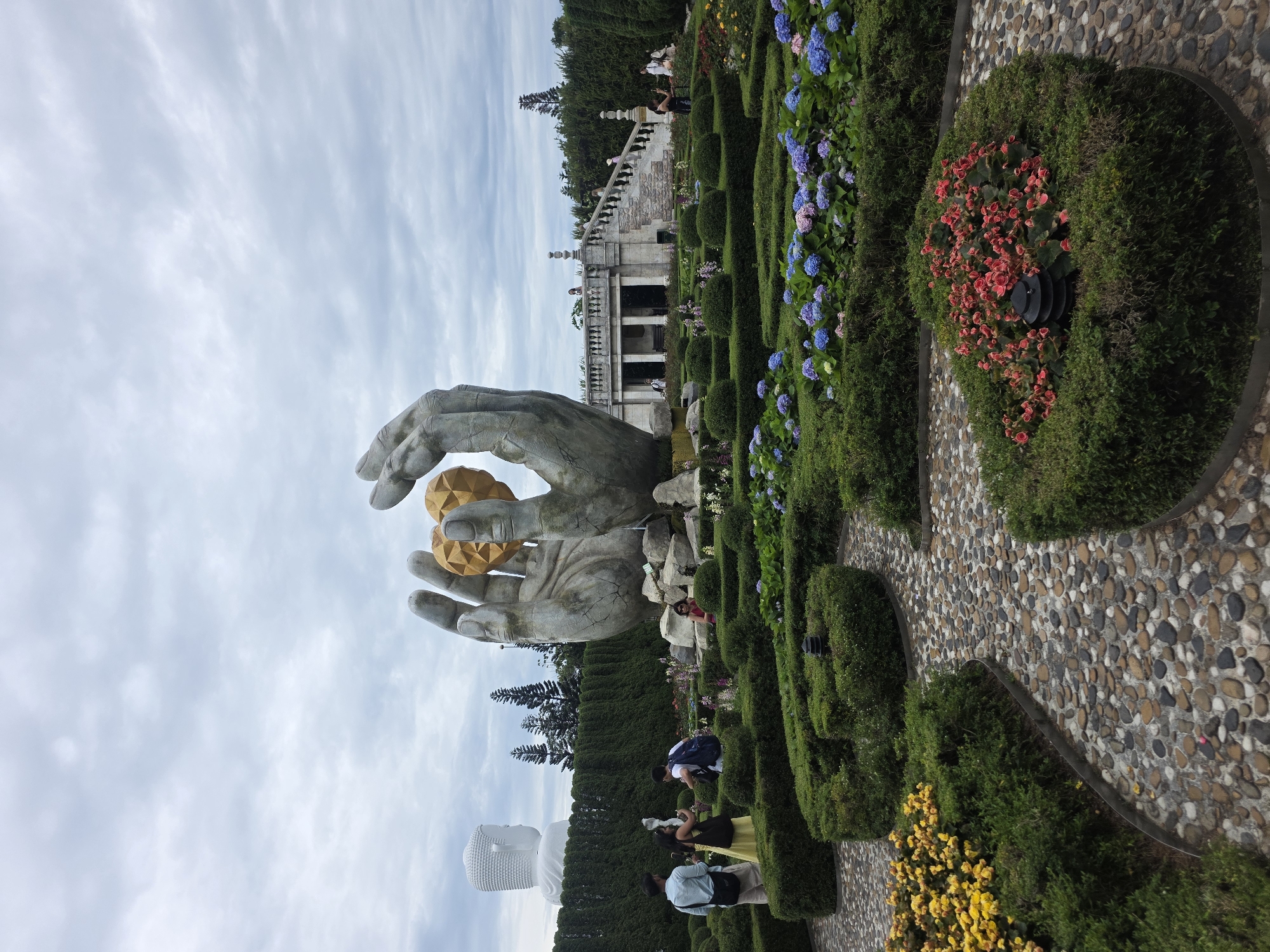 This image depicts a landscaped garden area with a prominent statue of two large, stone hands holding a golden sphere. In the background, a large white Buddha statue is visible, as is a white building with columns and a staircase. The garden features manicured hedges, cobblestone pathways, and colorful flower beds, including yellow, pink, and blue hydrangeas. Several people are present in the scene, some walking and others observing the statues and garden. The sky is overcast. This is the Ba Na Hills Golden Bridge in the city of Đà Nẵng, Vietnam.