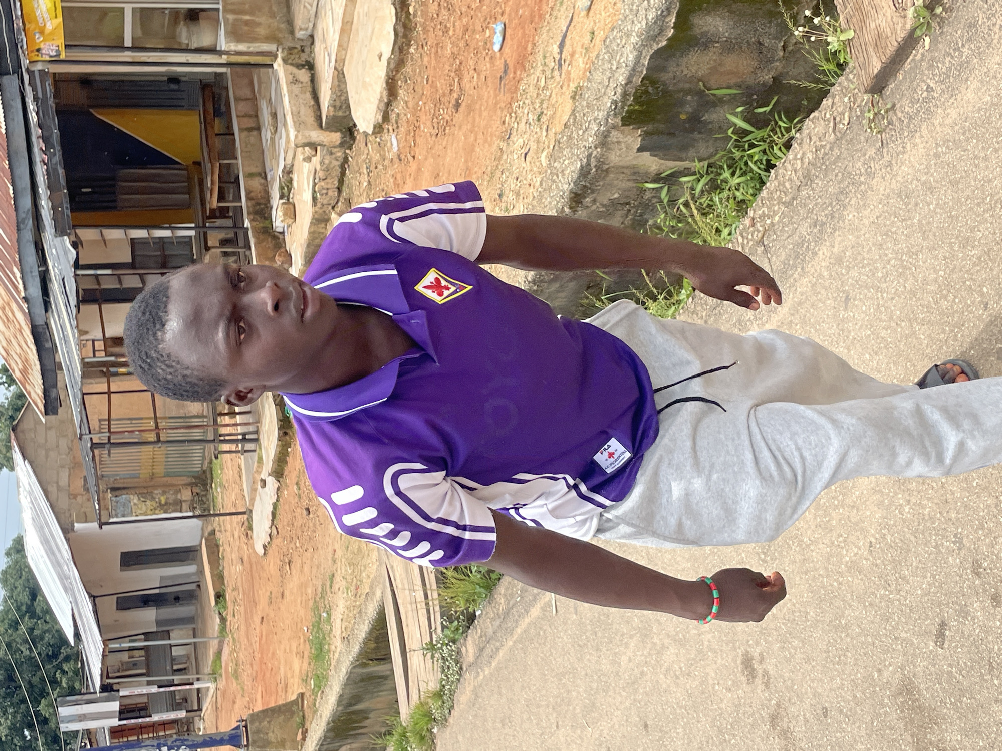 A young Black male is walking on a concrete path in Jos, Nigeria. He is wearing a purple polo shirt with white detailing and light gray sweatpants. He has a red, green, and black beaded bracelet on his left wrist. The background shows buildings with corrugated metal roofs, some appearing weathered, and a dirt road. The path the man is walking on has a concrete curb along the side, with some greenery growing alongside it. The overall scene depicts a casual outdoor environment within a residential or commercial area of the city.
