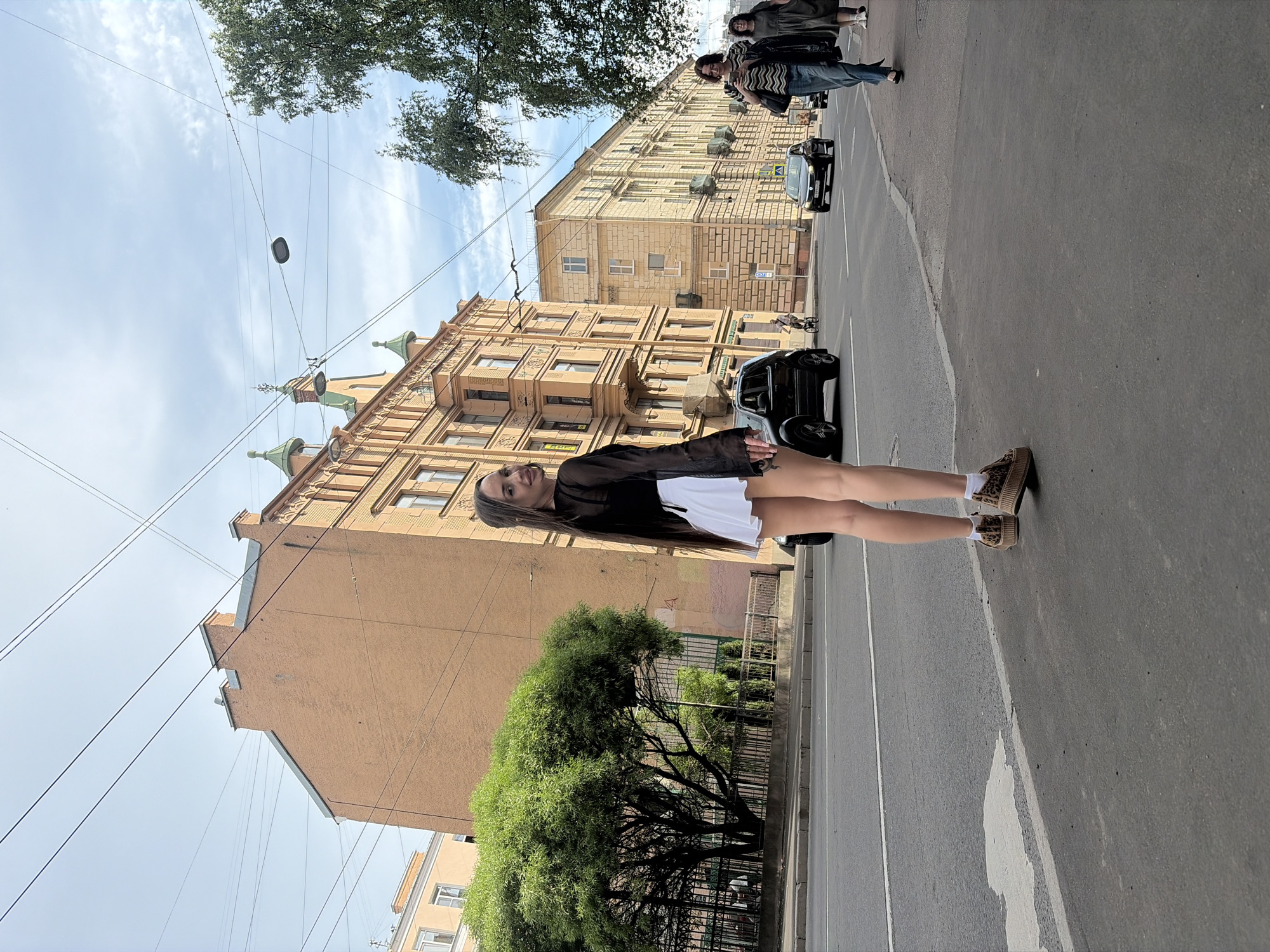 This image depicts a street scene in Saint Petersburg, Russia. In the foreground, a young woman with long, dark hair stands on a paved sidewalk, positioned with her back partially to the camera but turning her head to her left to look directly at the viewer with a slight smile. She is wearing a sheer black long-sleeved top over a dark undergarment, a white pleated mini-skirt, white socks, and platform shoes with an animal print.
Behind the woman, an asphalt road with a solid white dividing line stretches horizontally across the mid-ground. A dark-colored SUV is partially visible on the street immediately behind her, followed by a dark sedan further down. A person on a bicycle is also discernible in the distance on the road. To the left, a large, dense green tree with a rounded canopy stands next to the road, behind which a black metal fence is visible. Further left, a prominent multi-story building of terracotta-orange color features ornate architectural details and distinct green dome-like spires along its roofline. Another lighter-colored building is partially visible on the far left.
On the right side of the image, the sidewalk extends, and two other women are walking away from the camera. The closer woman wears a striped top and blue jeans, holding a dark bag and looking at a mobile phone. The second woman, further back, wears a dark dress. A large tree with green foliage hangs into the frame from the upper right. Lining the right side of the street is a long, multi-story, light-colored building with numerous windows and decorative architectural elements.
The sky above is partially cloudy, showing patches of blue. Numerous overhead utility wires or tram lines crisscross the sky, from which a single modern street lamp fixture is suspended.