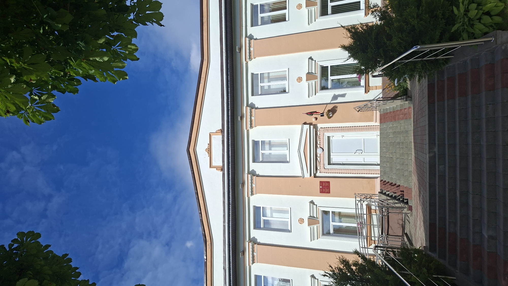 The image shows the entrance to a building with a blue sky and green tree leaves framing the top. The building has a light-colored facade with pale peach-colored vertical accents and decorative columns. There is a wide set of stairs leading up to a white double door. The stairs are made of gray and red paving stones. To the left of the stairs, there is a ramp with handrails. To the right of the stairs, there is a small evergreen tree and some low-lying green plants. A small red sign with text in a Cyrillic alphabet is visible to the left of the main entrance. A flagpole with a red and white flag is mounted on the wall to the right of the entrance. The building appears to be a public institution, potentially a government office or school, given the signage and architectural style. The location is identified as the Myadzyel District Executive Committee building in Myadzyel, Belarus.