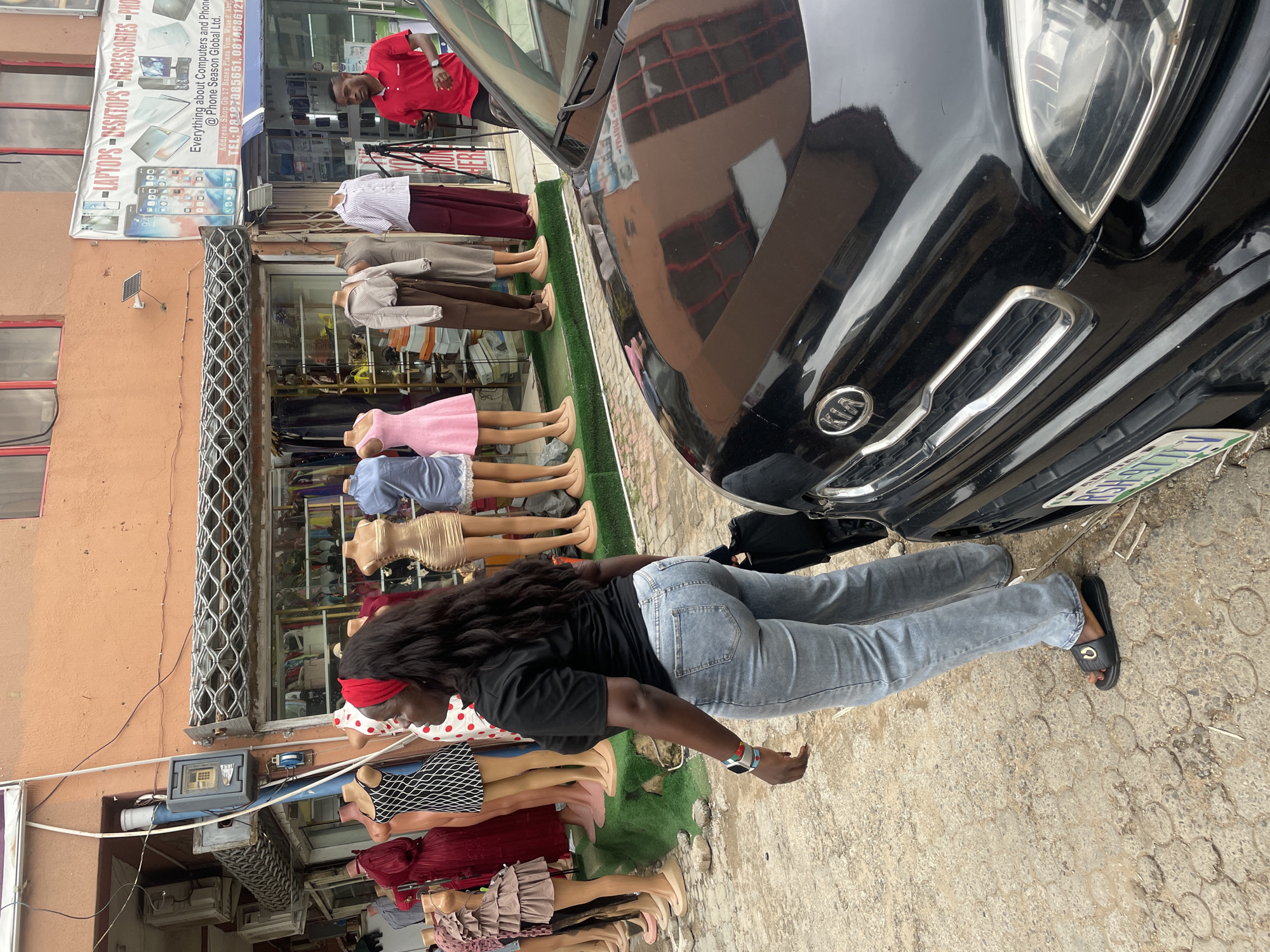 The image depicts a street scene in Abuja, Nigeria, with a focus on a woman standing next to a black Kia car. The woman, wearing light blue jeans, a black t-shirt, and a red headband, is seen from the side, looking towards a storefront. The storefront displays mannequins dressed in various female clothing items, including dresses and skirts. Above the storefront, a banner advertises "LAPTOPS - DESKTOPS - ACCESSORIES - PHONES" from "Phone Season Global Ltd." with an address in Banex Plaza, Wuse 2. The environment appears to be a commercial area, with visible air conditioning units and electrical conduits on the building. The car's hood and headlight are prominent in the foreground. A man in a red polo shirt is visible to the right, near the storefront. The ground is paved with interlocking stones.