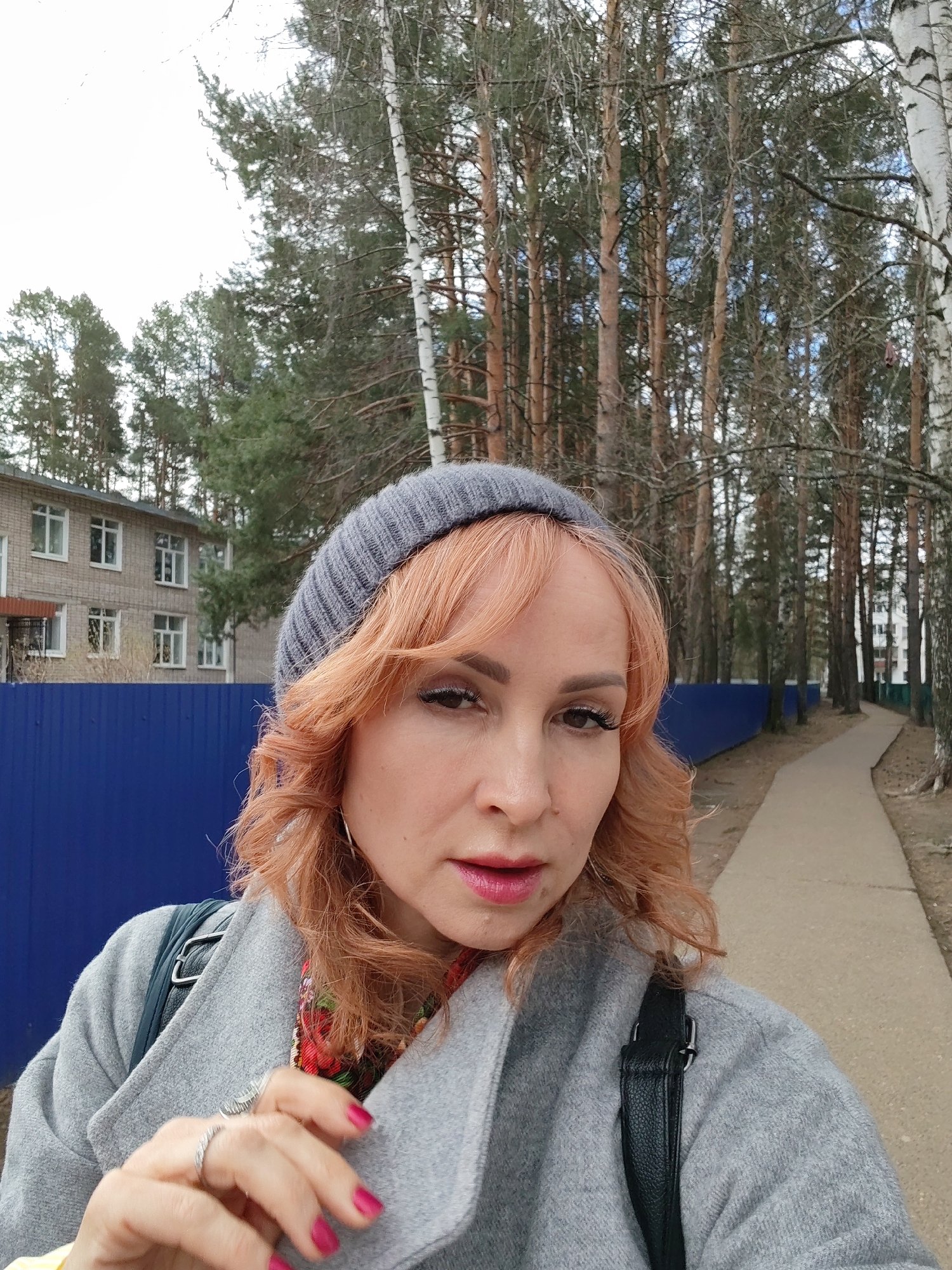 A selfie of a woman with fair skin, shoulder-length wavy orange hair, wearing a gray knit cap and a gray overcoat is set against a backdrop containing both urban and natural elements. A building with light gray siding and white-framed windows is visible behind a dark blue fence, suggesting a residential or institutional structure. A narrow concrete path extends into a dense, coniferous forest with slender birch trees. The location is likely in Chaikovsky, Russia.
