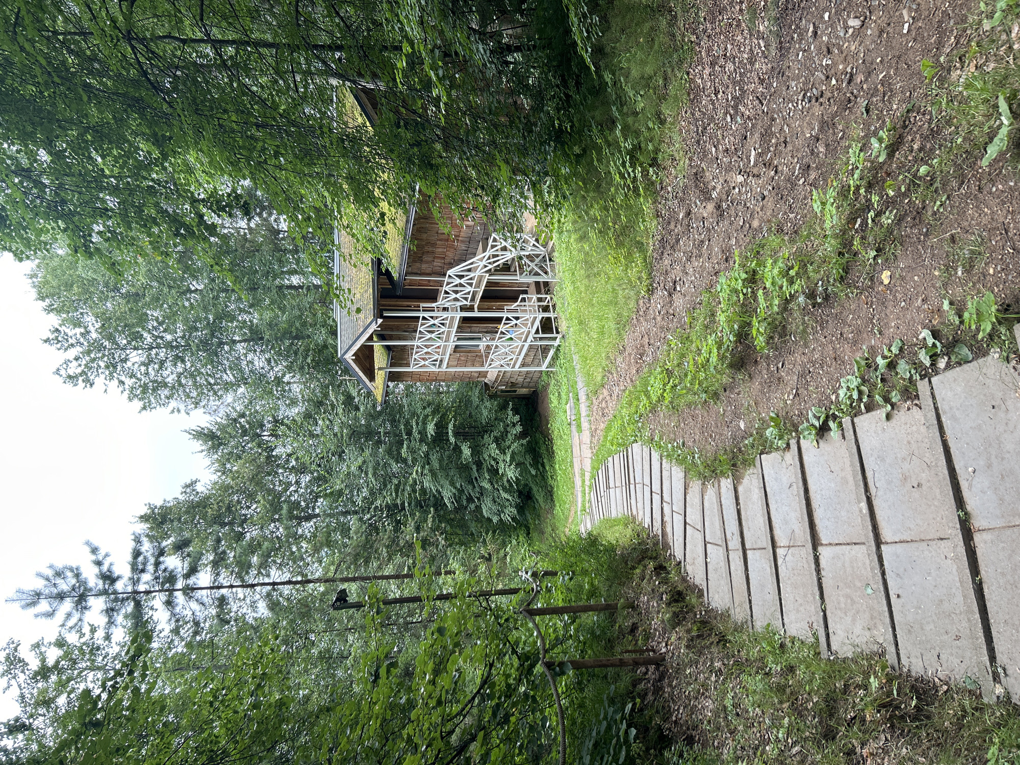 An overhead, slightly angled medium shot captures a winding stone pathway descending through a lush, green forest towards a rustic wooden building in Kostovaty, Russia. The path is constructed of rectangular, flat stones laid out in an offset pattern, creating a gentle curve that leads the eye downward and to the right. The stones are grey and appear weathered, with small patches of green vegetation growing in the cracks between them and along the edges of the path.
To the left of the path, a dark wooden post with a wire rope railing runs along the slope, disappearing into the dense foliage. The forest is characterized by tall, mature trees with thick green leaves, creating a canopy that filters the daylight and lends a serene, natural ambiance to the scene. In the background, to the right of the path, stands a two-story wooden house. The house has a brown, shingled exterior and a moss-covered green roof. It features white, decorative railings and staircases on its exterior, suggesting a traditional architectural style. The building is partially obscured by the dense trees and branches that frame the right side of the image. The ground surrounding the path and house is covered with dirt, fallen leaves, and patches of grass and low-lying plants. The sky is overcast and a pale grey, indicating a cloudy day.