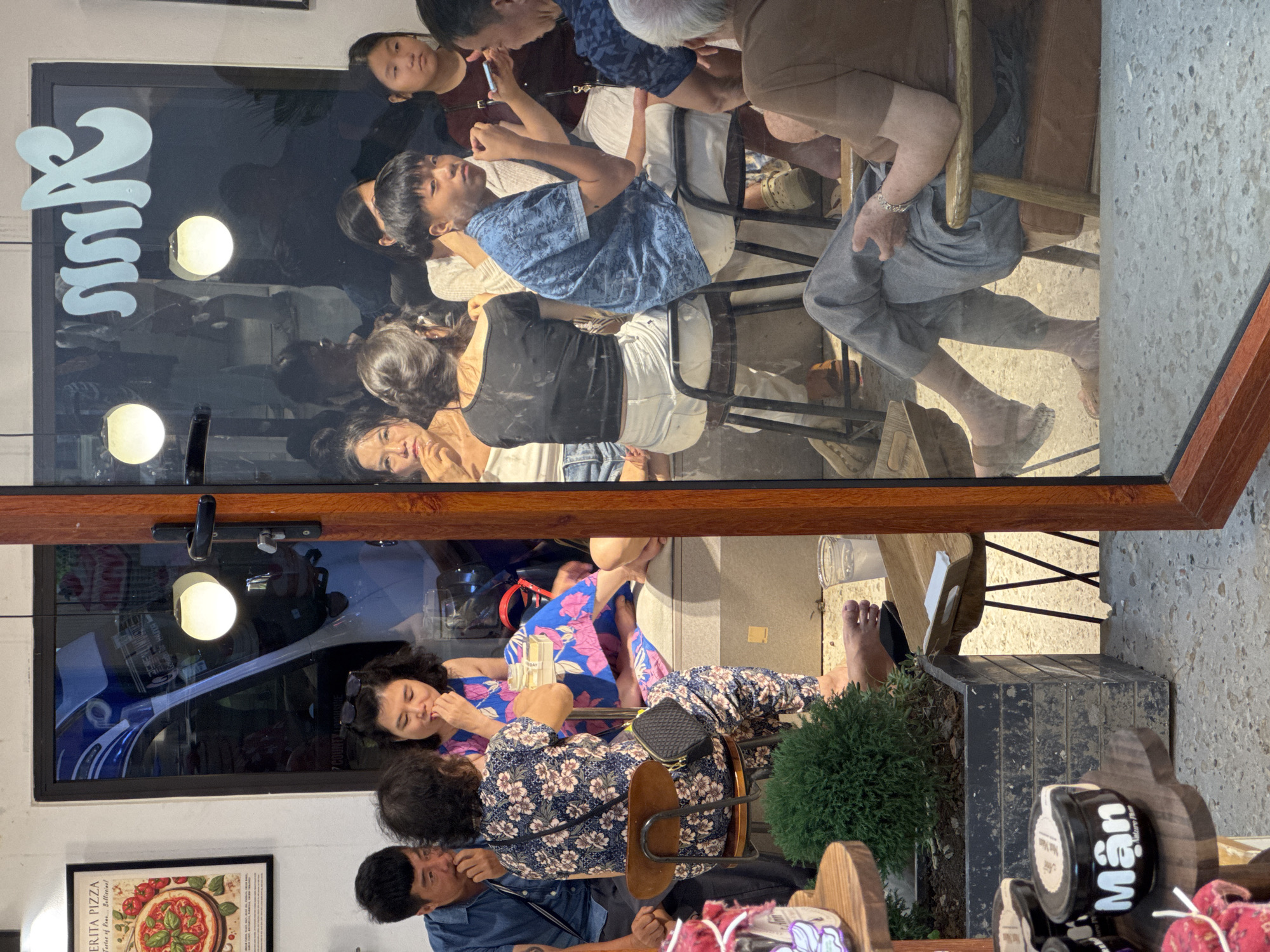 The image captures a group of people inside a cafe in Mộc Châu, Vietnam, viewed through a large glass window. In the foreground, on the left, a wooden shelf displays several small jars, one of which is labeled "Mân". To the right of the shelf is a black planter containing a small green bush. A clear plastic cup with a light-colored liquid sits on the floor near the planter.
Inside the cafe, several individuals are seated at tables. On the left side of the cafe, a man in a blue shirt is seated next to a woman in a floral print outfit. The woman is seated on a stool and appears to be eating. Further back, a woman with dark hair is also seated.
In the center and right side of the cafe, more people are visible. A young man in a blue patterned shirt is seated on a stool, looking towards the camera. A young woman is seated next to him, wearing a black off-the-shoulder top and white pants. Other individuals are partially visible in the background, including a woman with dark hair on the far right. The cafe interior is dimly lit with several round hanging lights. The reflection on the glass shows some signage with stylized text. The overall scene suggests a casual gathering in a cafe setting.